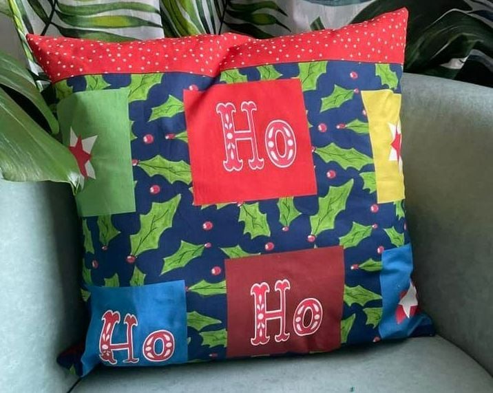 Christmas cushion in dark blue with green holly leaves and red berries. There are large coloured squares with Ho on each