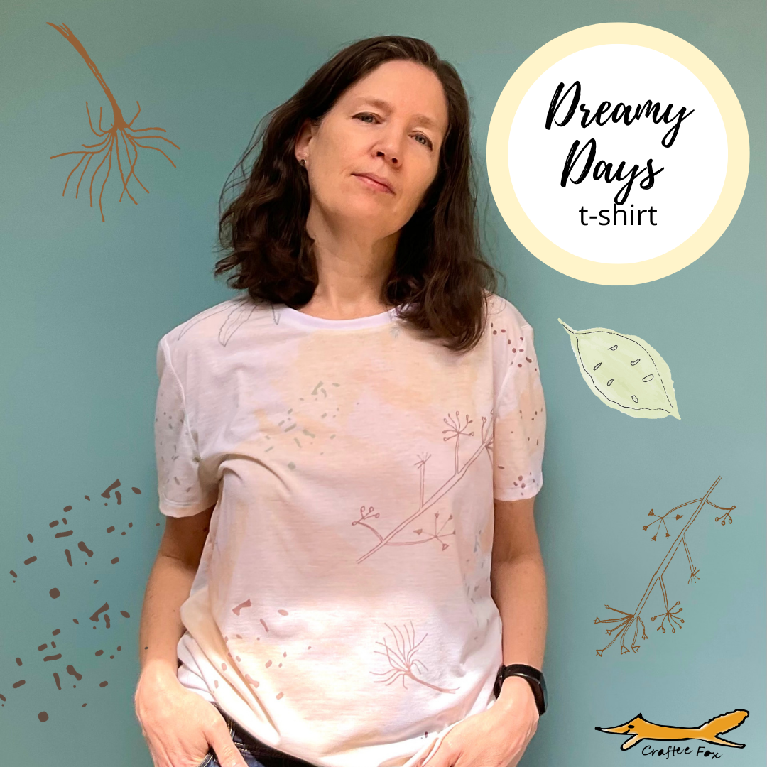 A 40 year old plus woman wear a pale yellow and cream t-shirt. The t-shirt has pale patches of blue and images of plants stalks and leaves are scattered around the t-shirt. A circle reads 'Dreamy Days' t-shirt
