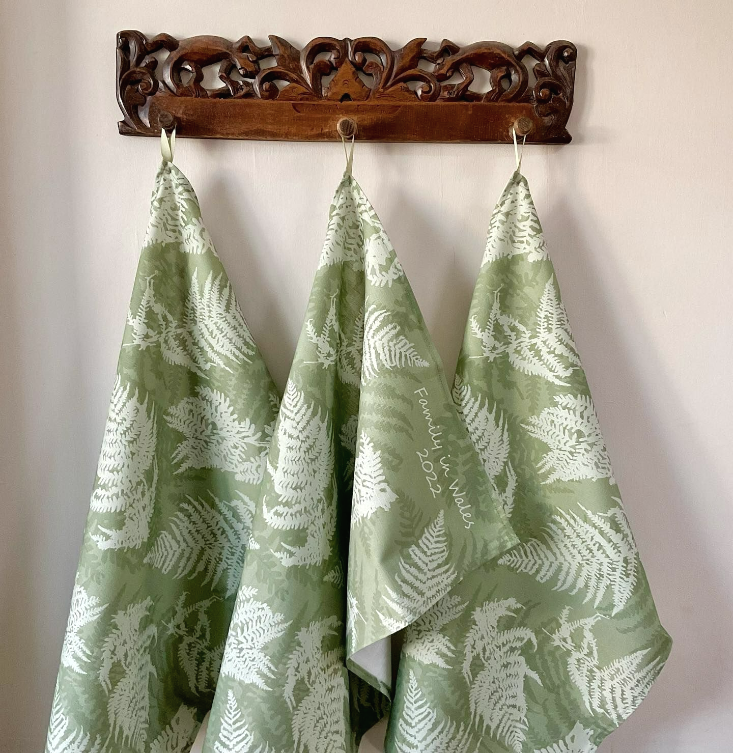Three green tea towels hang off pegs. They are green with a fern leaf print