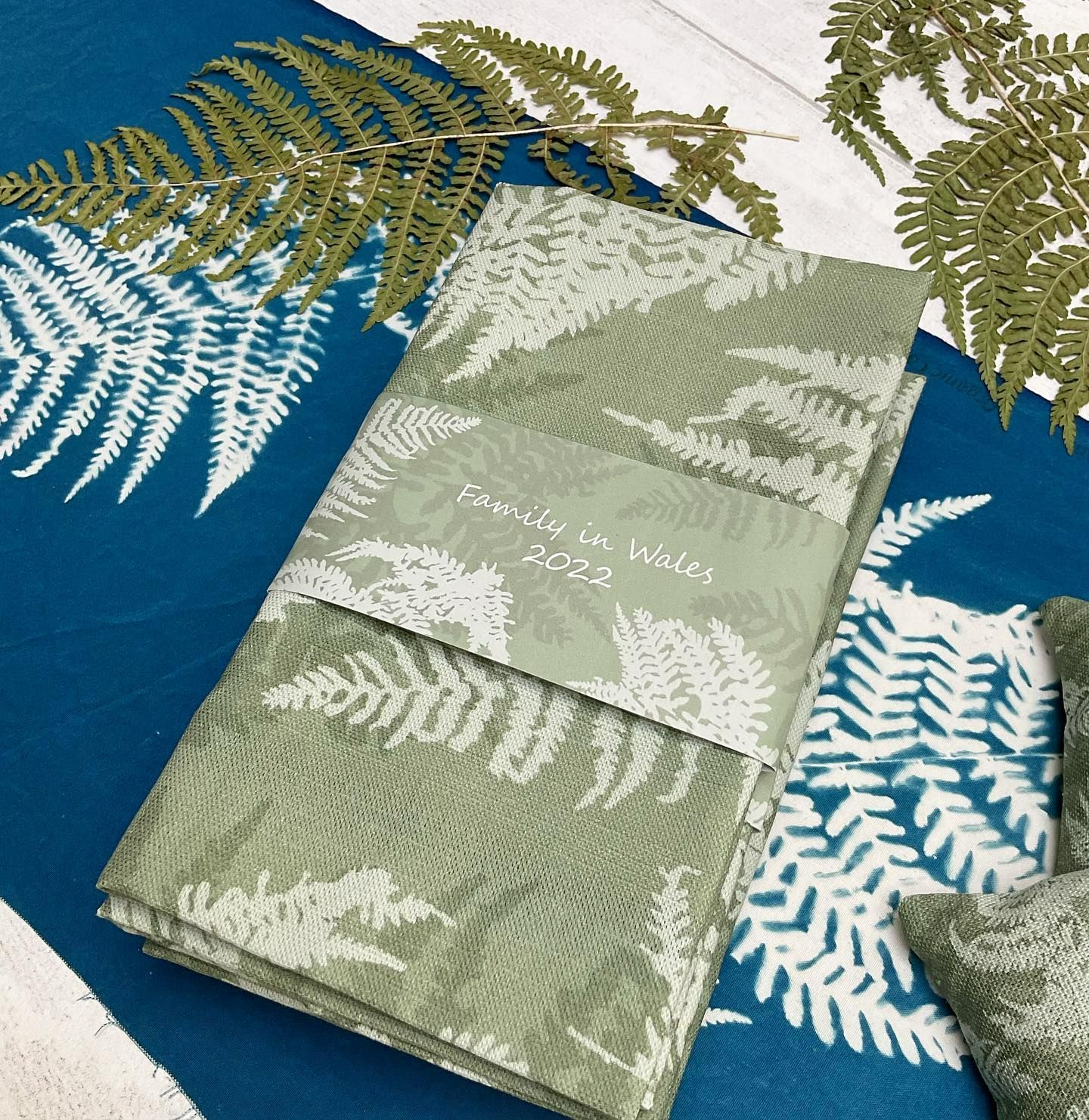 A green tea towels with fern print is folder on a blue background.