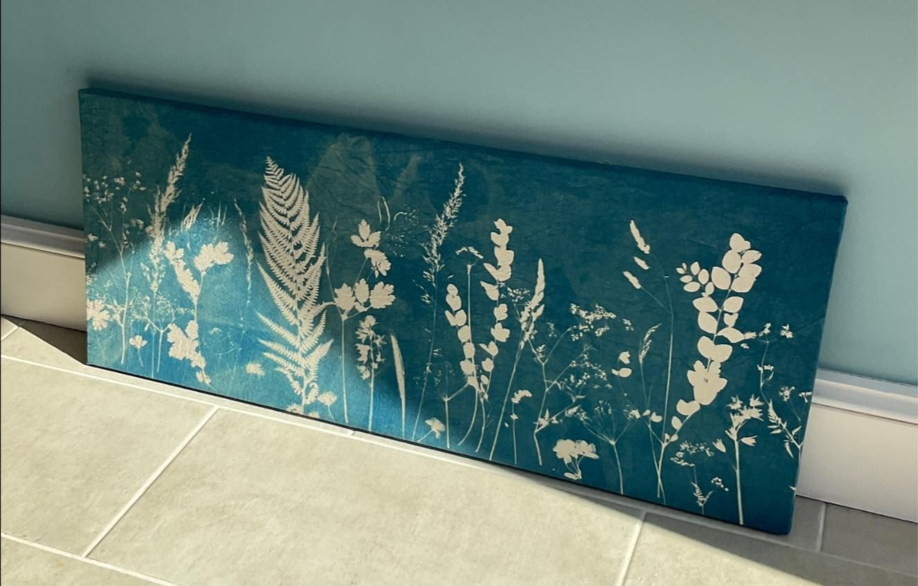 A large sunprinted cyanotype of hedgerow plants and Agapanthus rests against a blue wall. The blue fabric is stretched over a wooden frame.