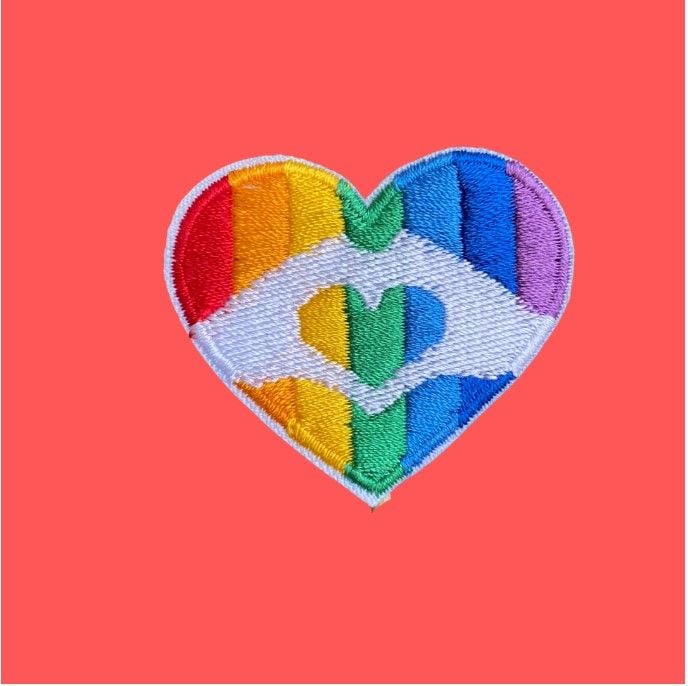 A heart shaped iron-on patch with a rainbow background and two hands forming a heart