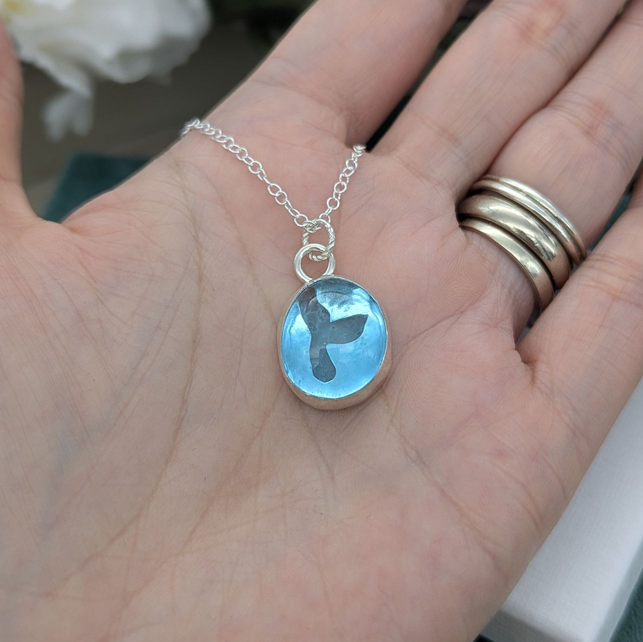 A aqua blue glass silver pebble necklace with a whale tail cut out detail being held in a hand