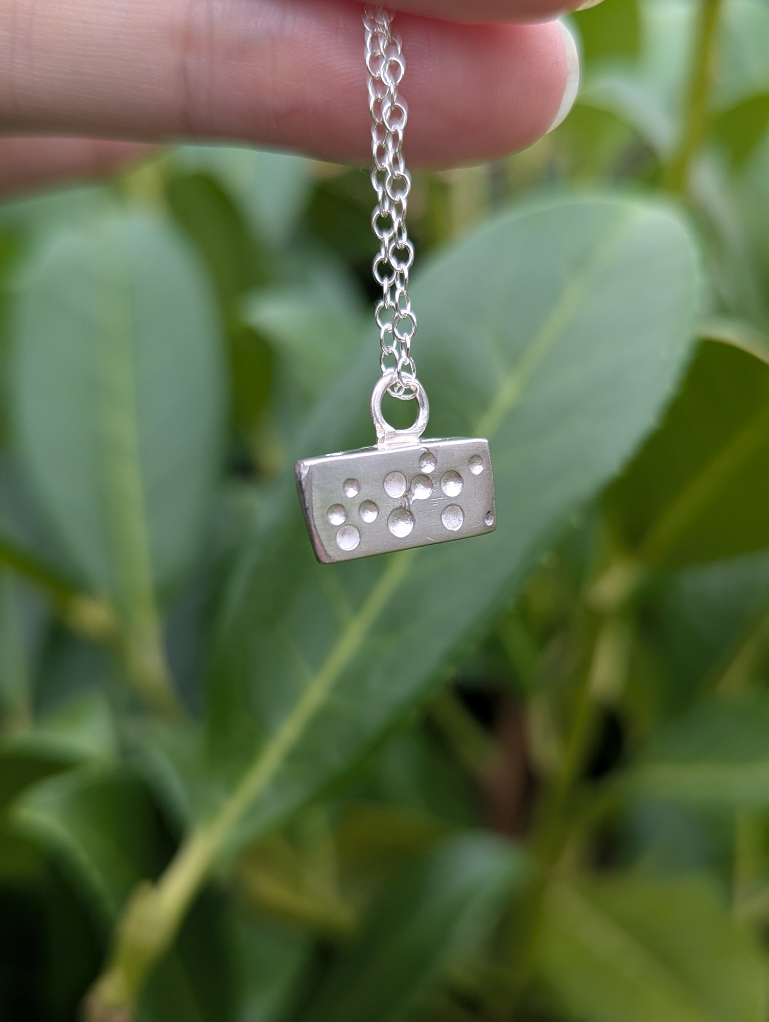 A sterling silver cheese shaped necklace hanging against a green leaf backdrop