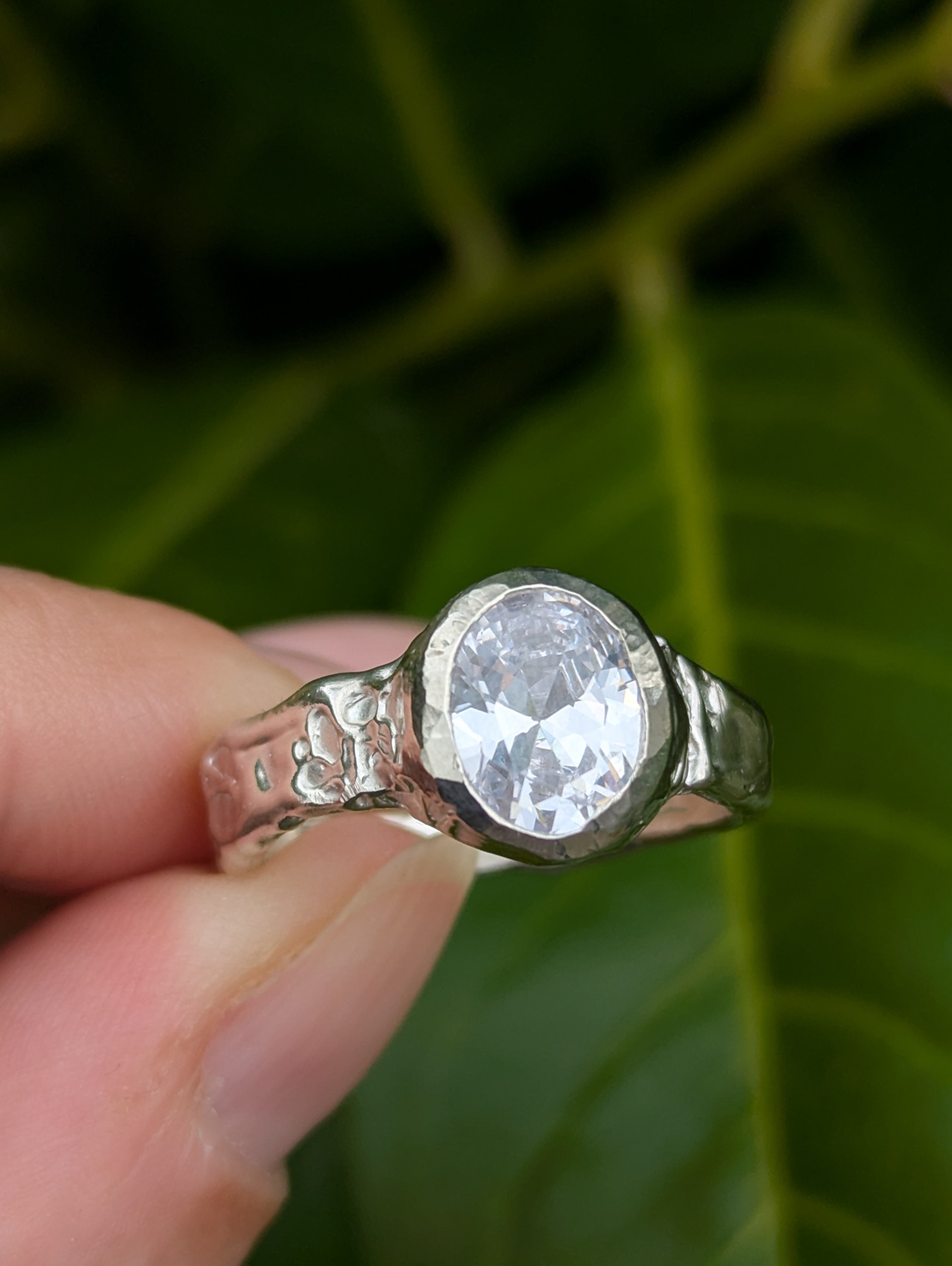 a unique silver ring set with a CZ facet cut gemstone being held in front of a leafy green back drop