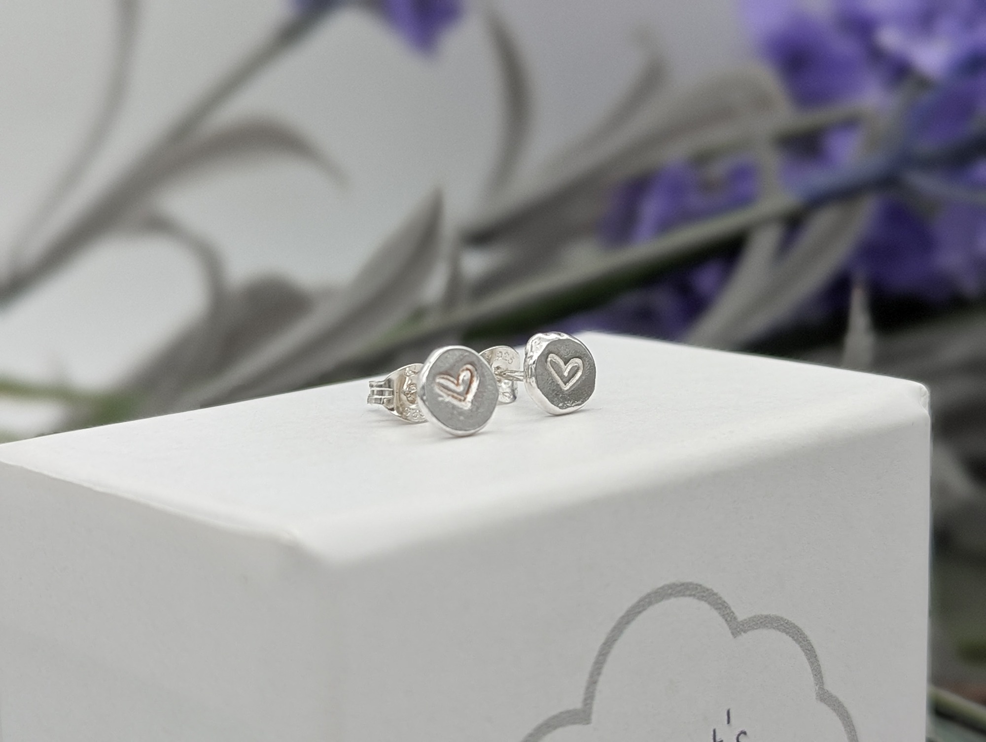Silver heart stamped stud earrings on a white jewellery box with a leafy background