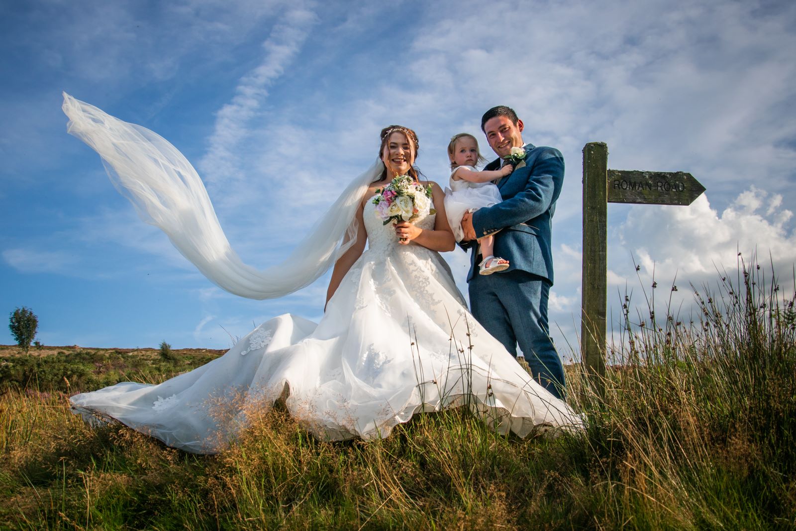 A bride, groom and their daughter stand on the moor and look down the camera while smiling. A gust of wind has come along and her veil is flying out to the left.