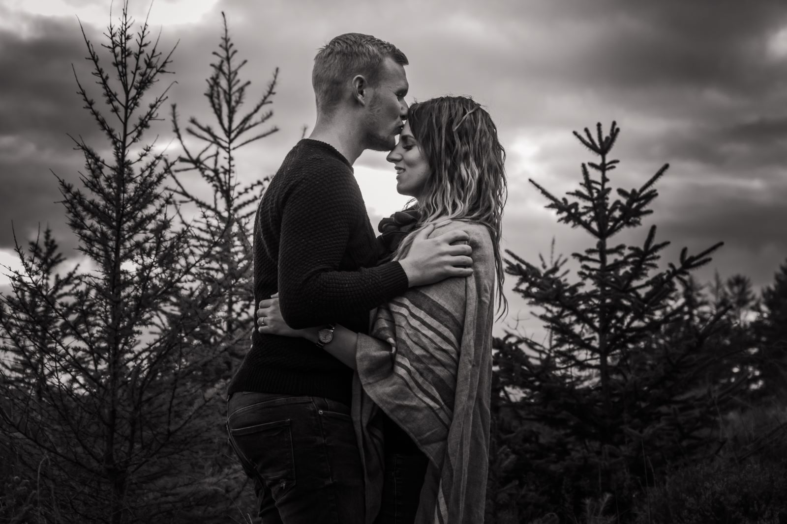 A groom-to-be is stood facing his fiance while kissing her forehead. There's christmas trees in the background and the image is black and white.
