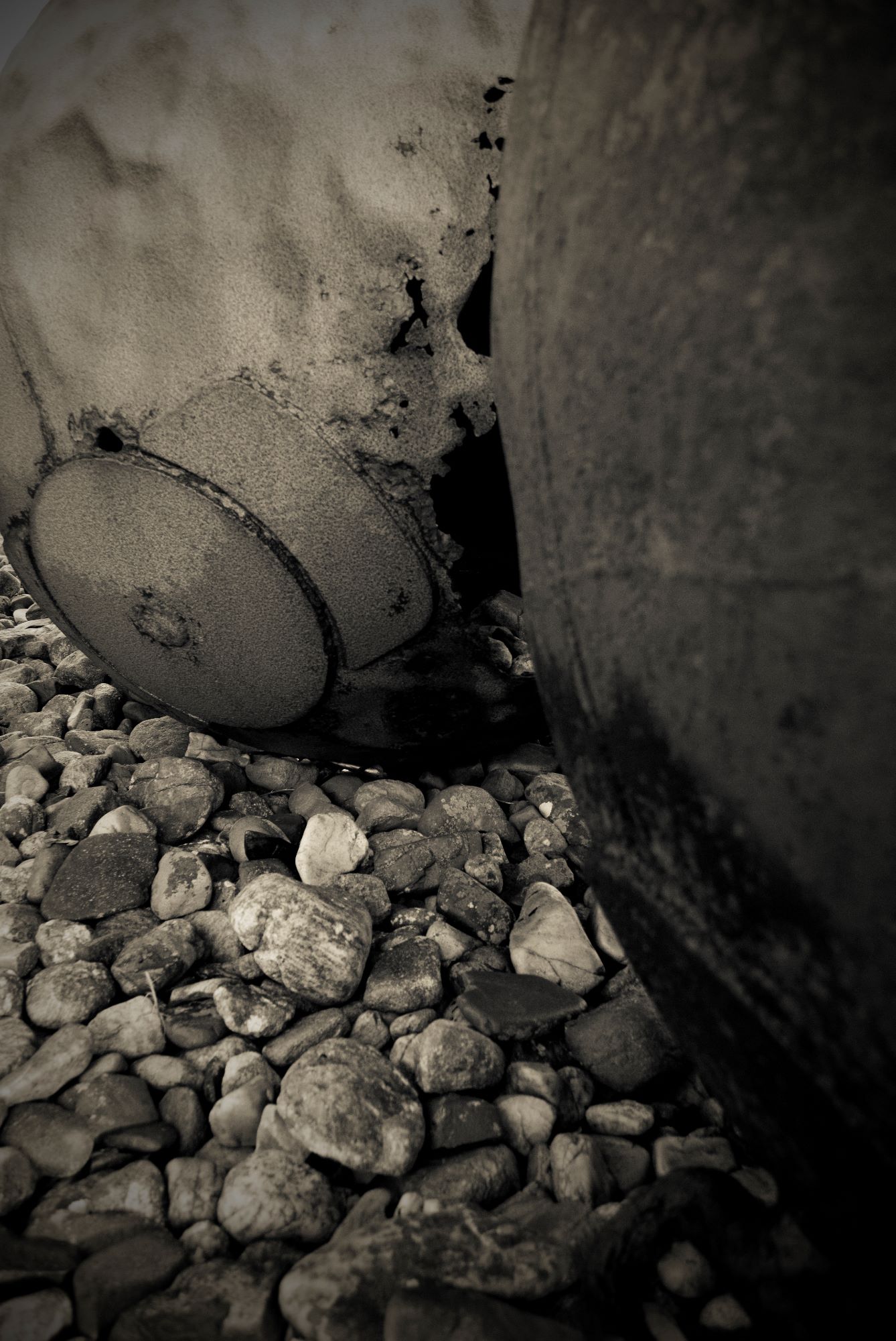 Old metal sea buoys sit on a pebbled beach as they slowly rust away. The image is black and white with lots of texture in.