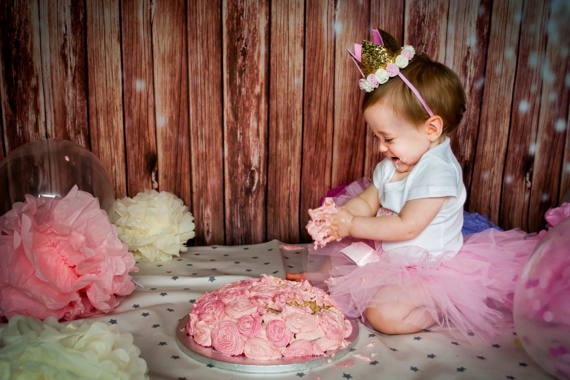 A little girl wears a white top and pink tutu and is sat by her pink birthday cake. She has her hands full of icing and is smushing it all over her fingers and arms