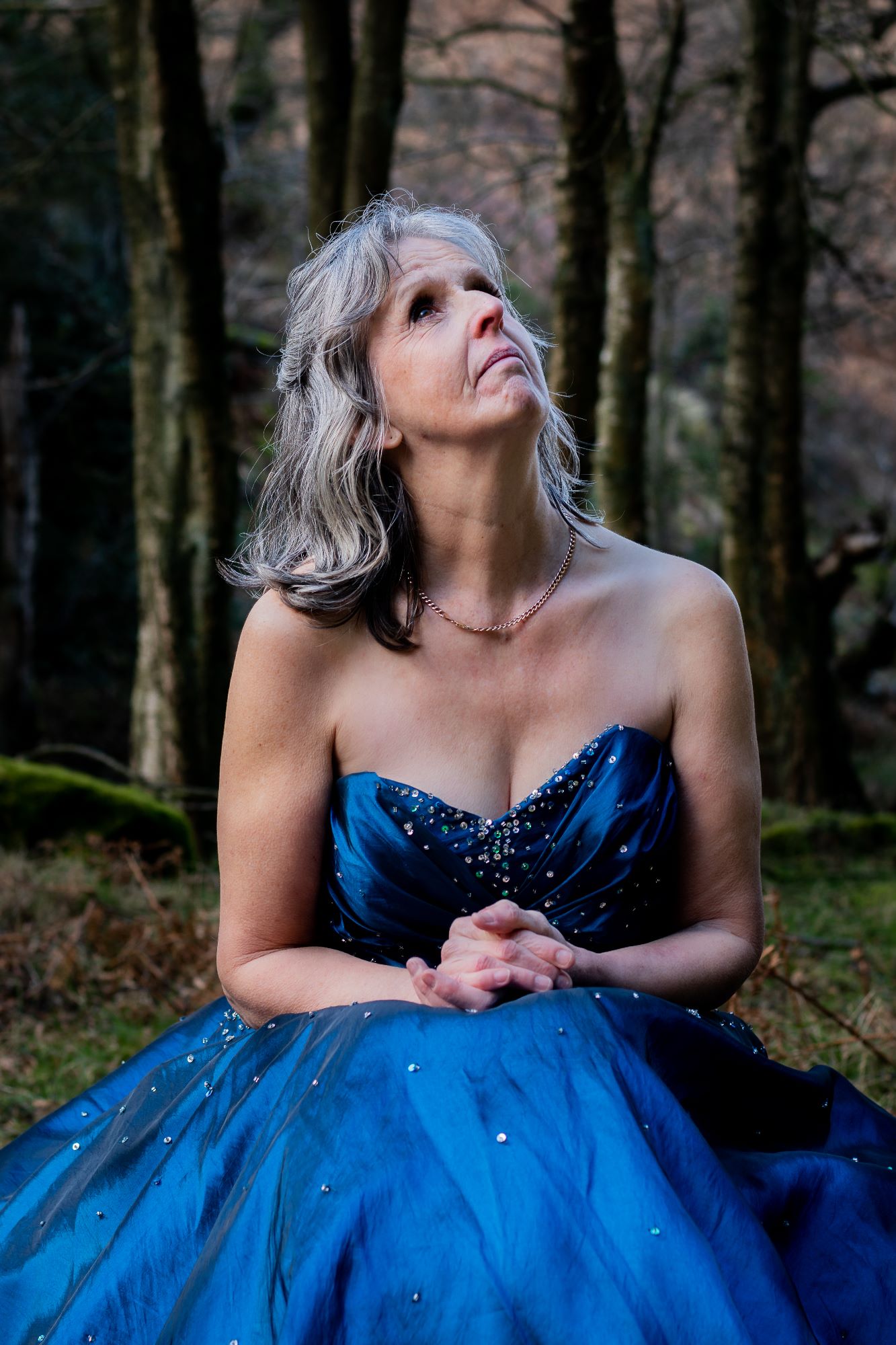A lady sits on a tree stump in the forest and looks towards the sky. She's wearing a blue ballgown and looks like a real life cinderella