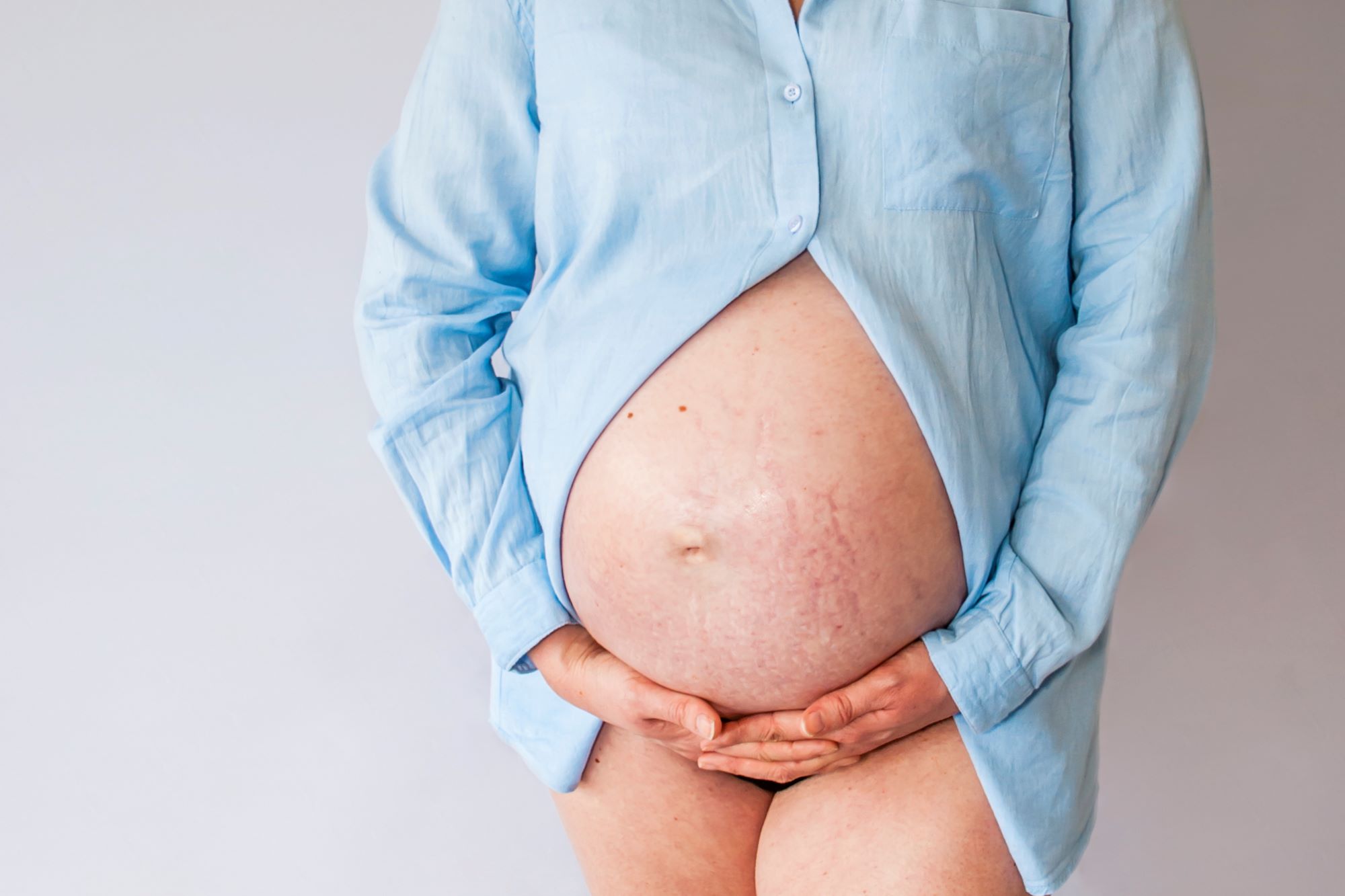 A mother to be  wears a blue shirt and cradles her bump straight on to the camera, her shirt is unbuttoned to show her bump fully