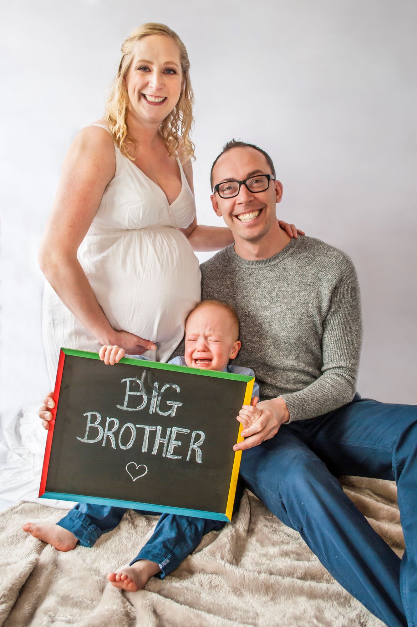 A little boy sits with his mum and dad and holds up a sign saying Big Brother. He's crying and mum and dad are smiling during their maternity photoshoot