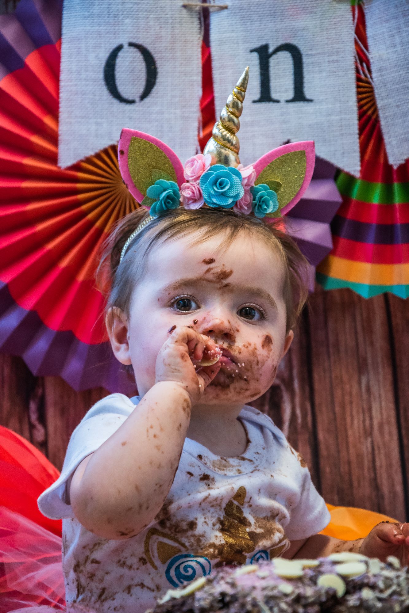 A little girl wears a unicorn crown and sits eating a chocolate button off her birthday cake. She has chocolate icing all over her face, arms and t-shirt which means it's been a successful cake smash!
