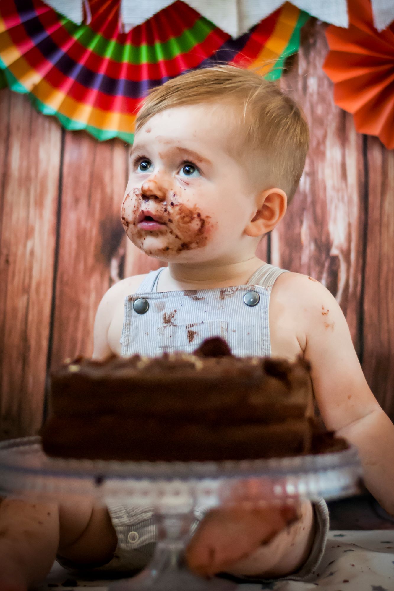 A little boy sits behind his chocolate cake and looks up at his mum and dad. He's got chocolate all over his face