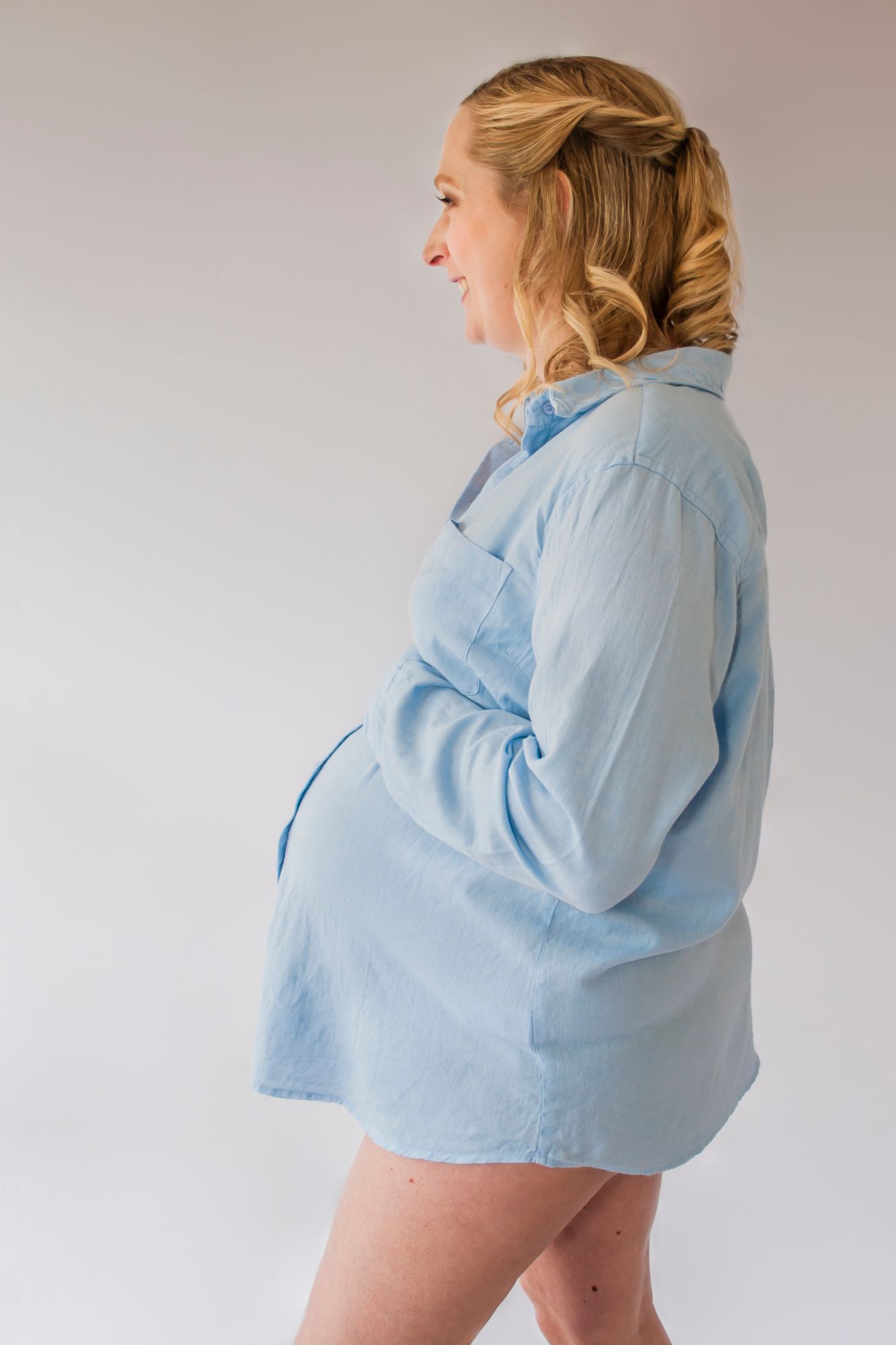 A mother to be stands in a blue shirt against the white wall in her kitchen and rests her hand on her bump