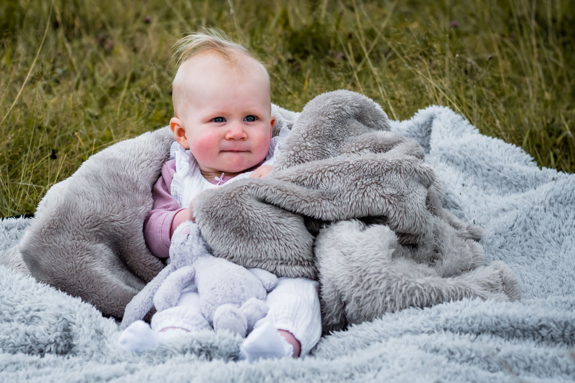 A little girl wraps herself in a blanket, the wind is blowing her hair and she has her bunny teddy on her legs. She's only six months old