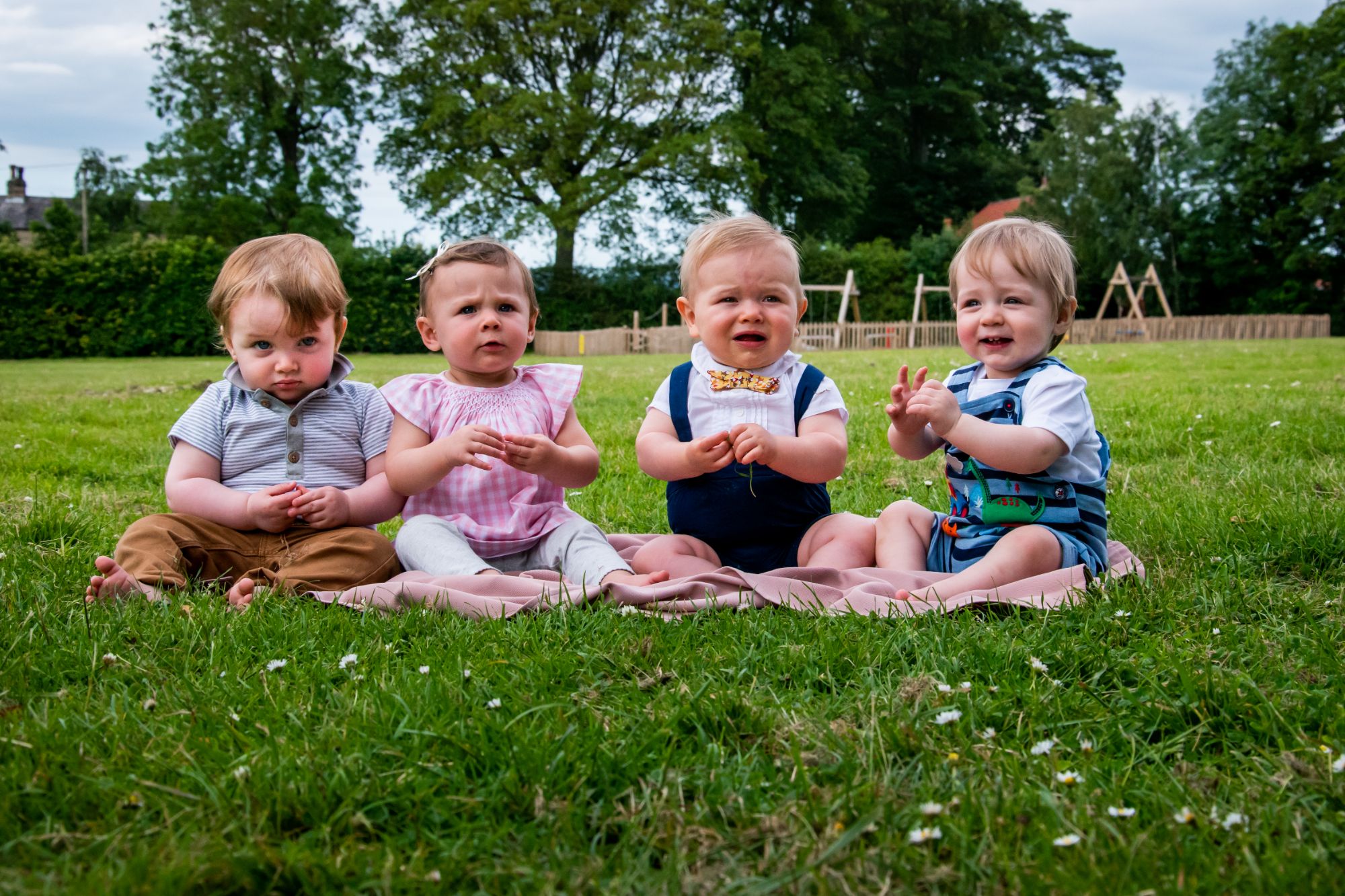 A little boy is joined by his one year old friends as the four of htem sit in a line outdoors at his first birthday party.