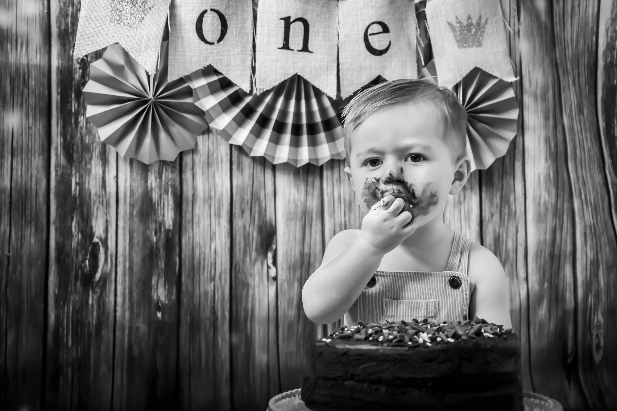 A little boy sits behind his cake and places the next bit in his mouth. A banner behind reads ONE and his face is covered in chocolate