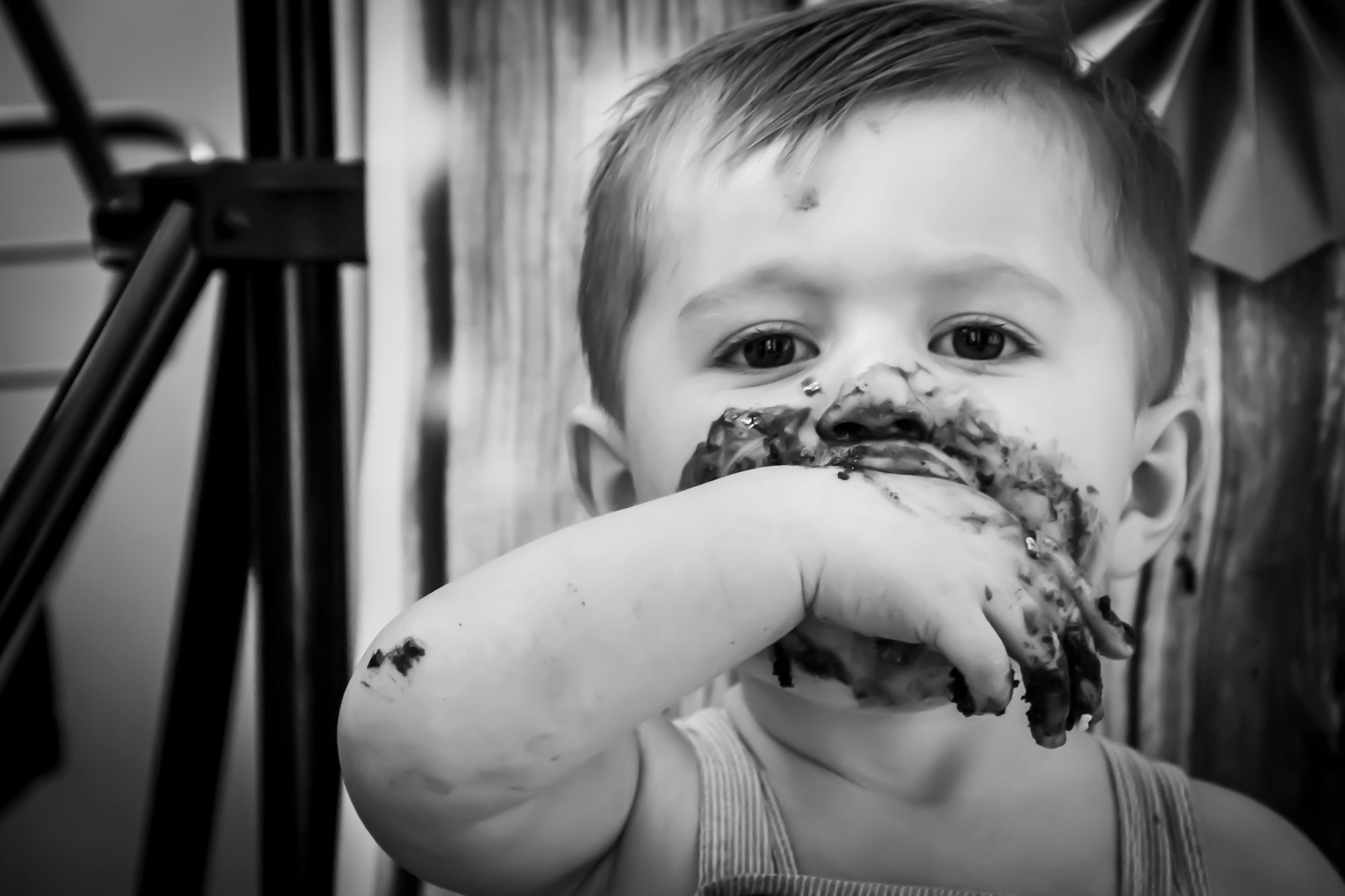 A little boy looks straight down the camera while sucking his thumb. His face is covered in chocolate icing but i