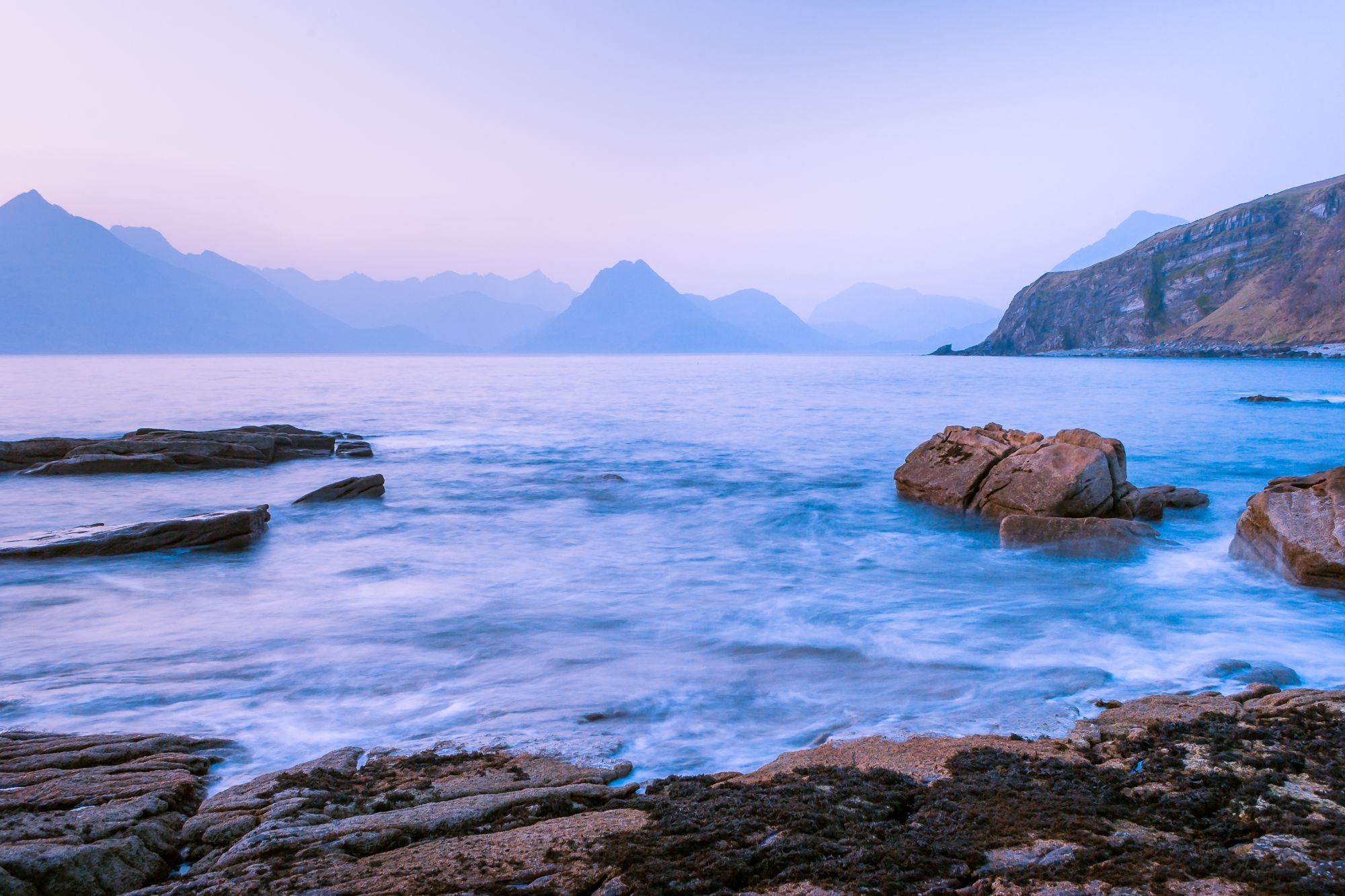 Elgol Beach at blue hour - the sea has been slowed down in camera to create a lovely swirling effect. The Cuillin mountains can be seen in the distance and there's a few rocks in the foreground