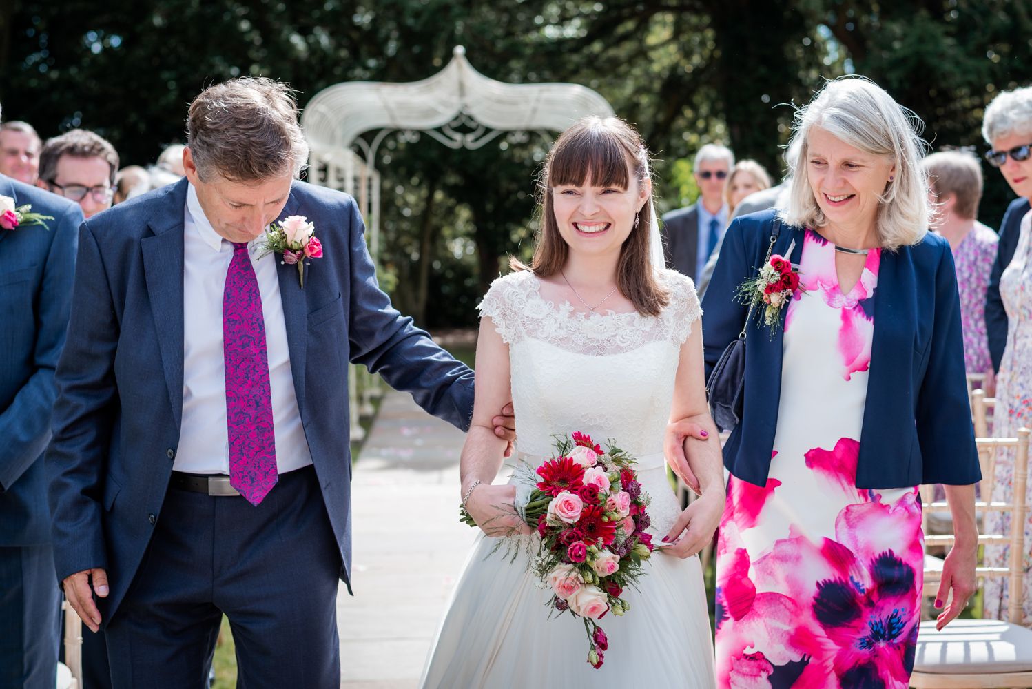 A Mother and Father walk their daughter down the Isle on her wedding day. The ladies are smiling and the father is looking down collecting his thoughts.