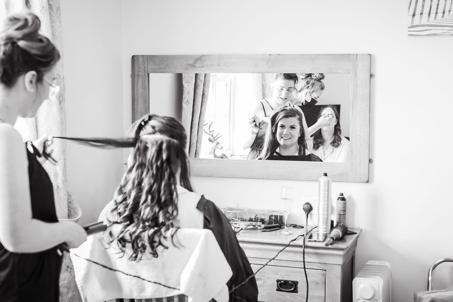 The bride is photographed from behind while she has her hair done on her wedding day. In the reflection of the mirror we can see her smiling face and her maid of honour having her make up done too.