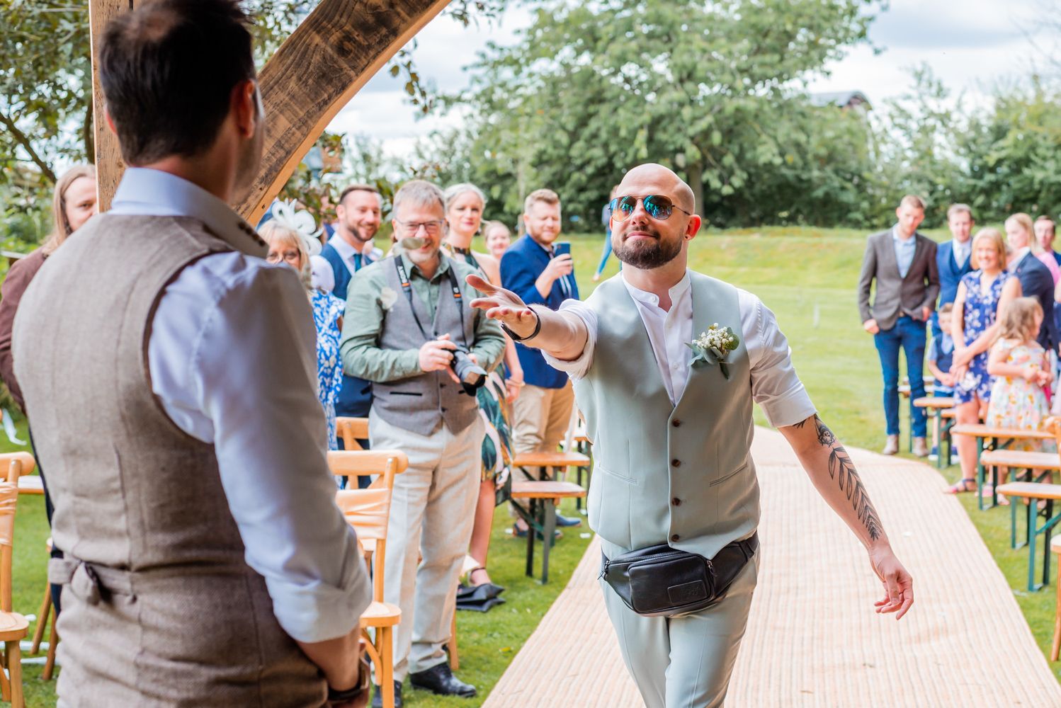 The bride's male best friend throws rose petals towards the groom after walking down the isle. The guests can be seen in the backgroud laughing.
