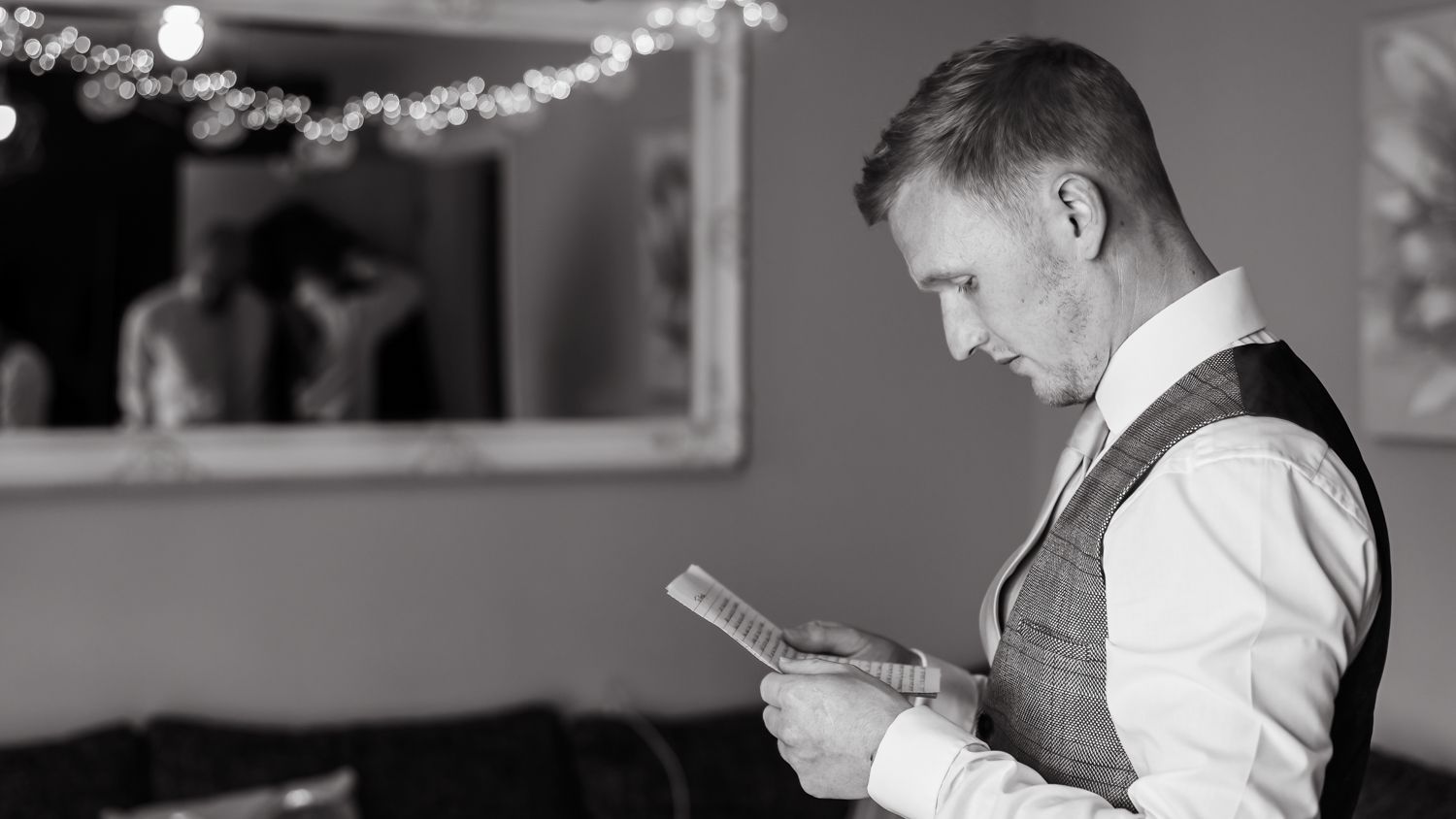 the groom reads a letter from his bride-to-be after getting ready on their wedding day. He's full of emotion and the groomsmen can be seen getting jackets reads in the reflection of the mirror.