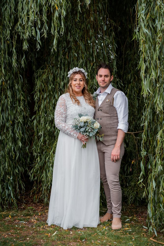 The bride and groom look towards tha camera as they stand under a willow tree for their portrait taken.