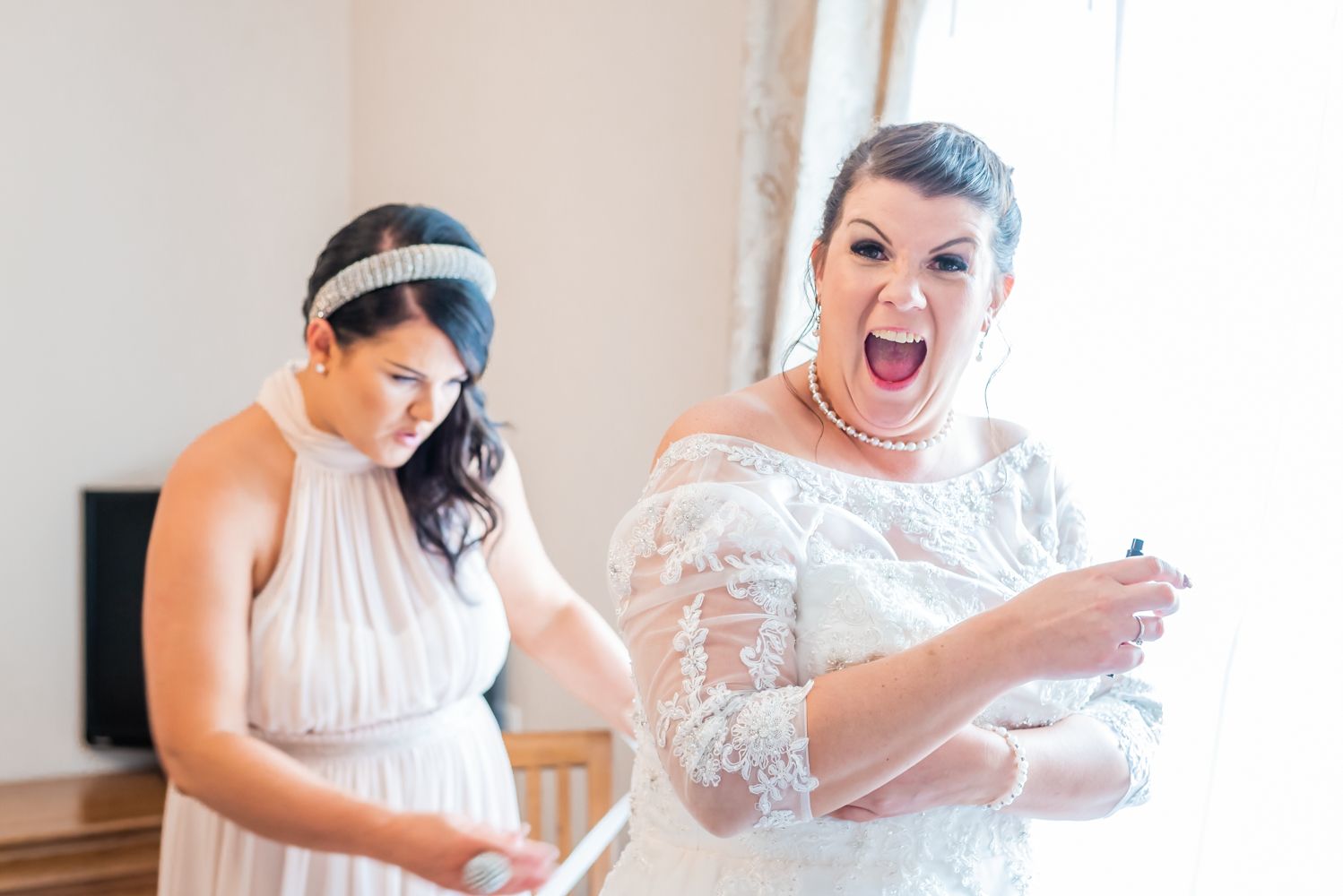The bride pulls an ecited face as she looks directly down the camera, her friend is doing up her wedding dress for her and she's excited to marry her man.