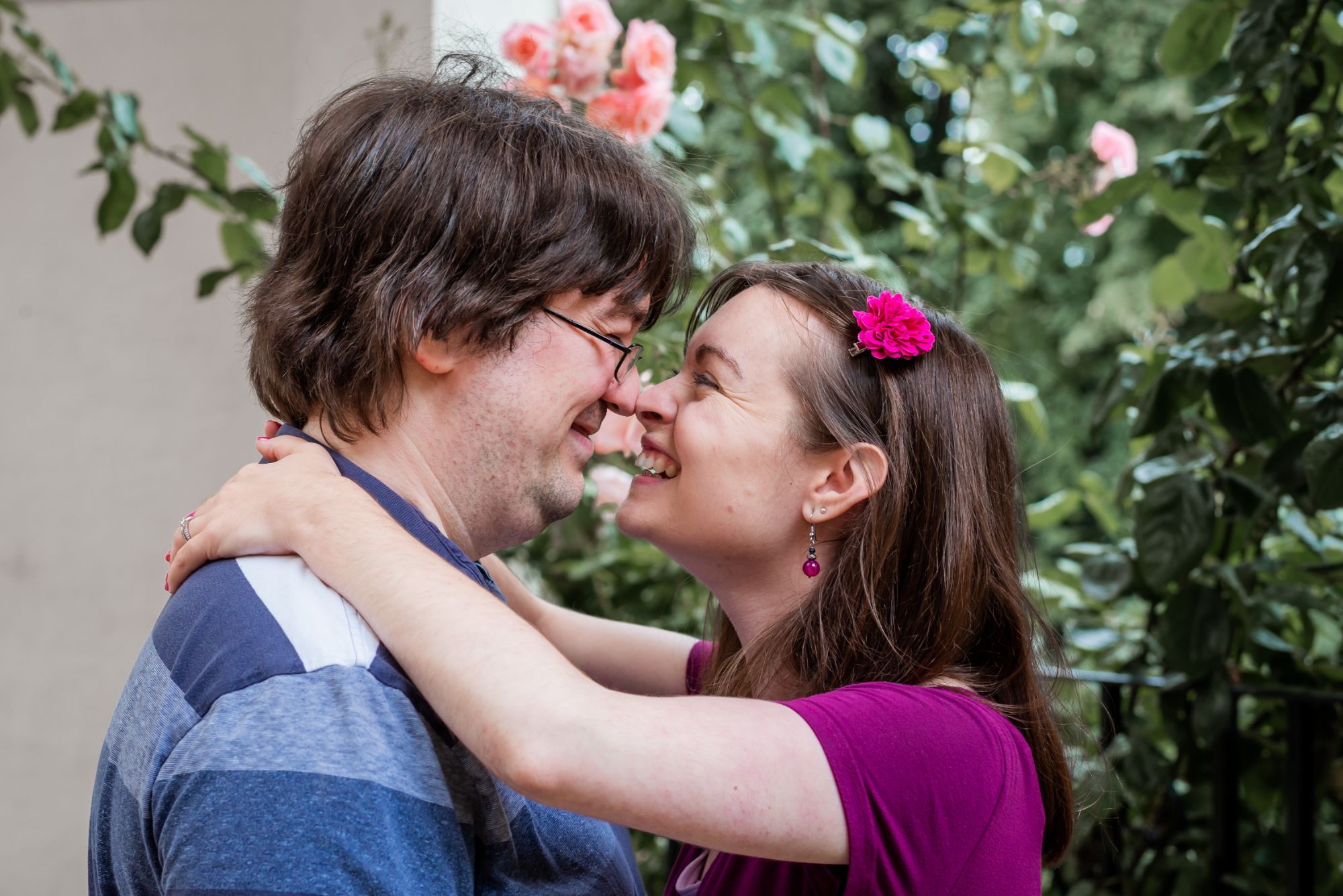 A man and lady touch noses and giggle at their engagement session. She has her arms wrapped around his shoulders and there's roses in the background