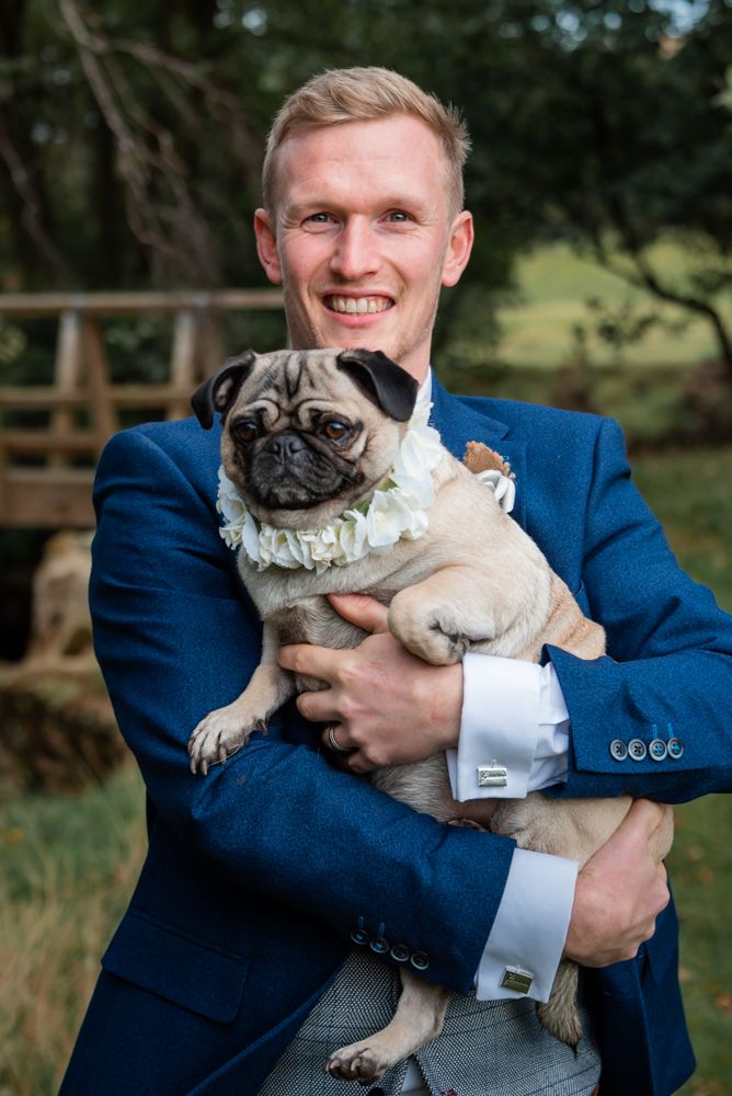 A Groom holds his pug while in his wedding suit and smiles as he looks down the camera.