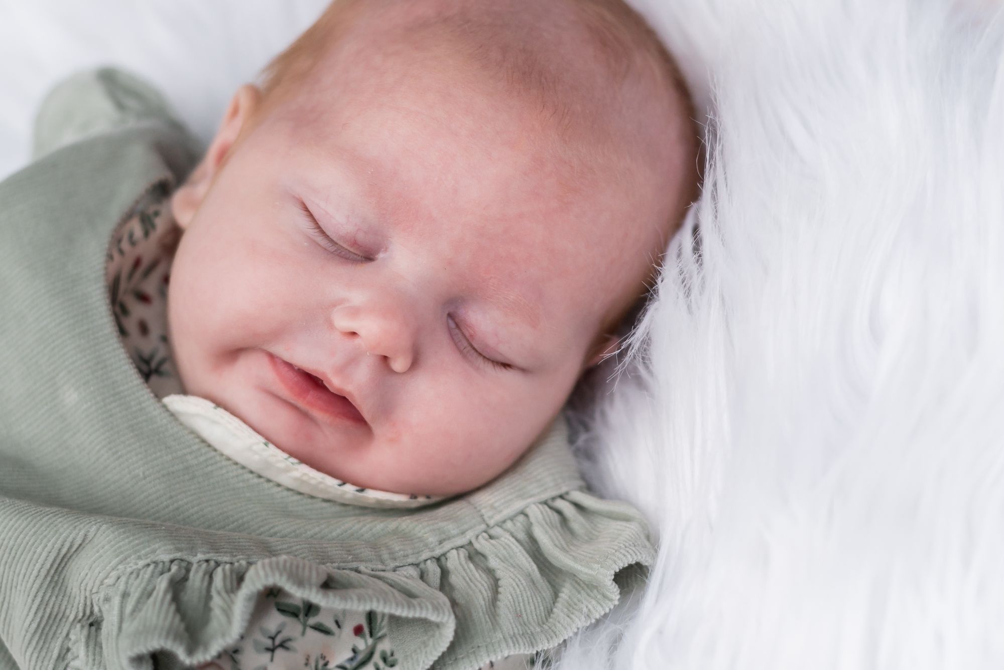 An eight week old baby at her newborn photoshoot lies on a fluffy white blanket and sleeps