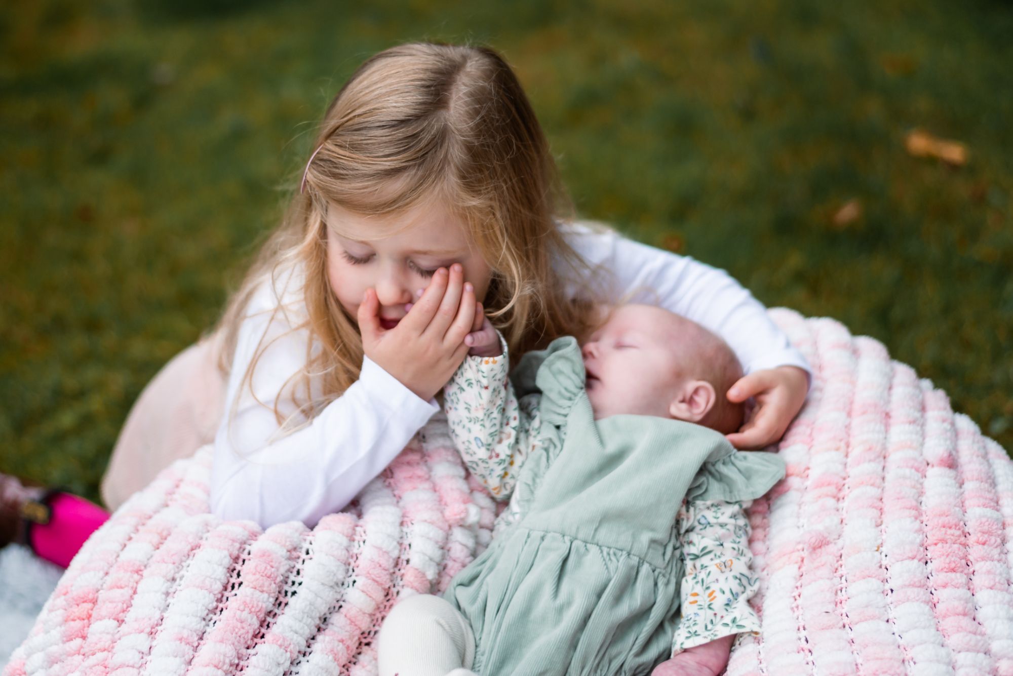 A big sister who's 4 is kneeling next to her new baby sister and is holding her tiny hand against her cheek. they both have their eyes closed