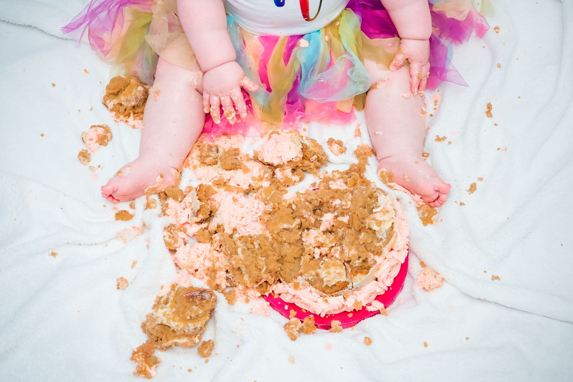 A cake is broken into tiny pieces and photographed from above. You can see two little legs and two little hands that are covered in icing after the little girl has enjoyed her cake smash session
