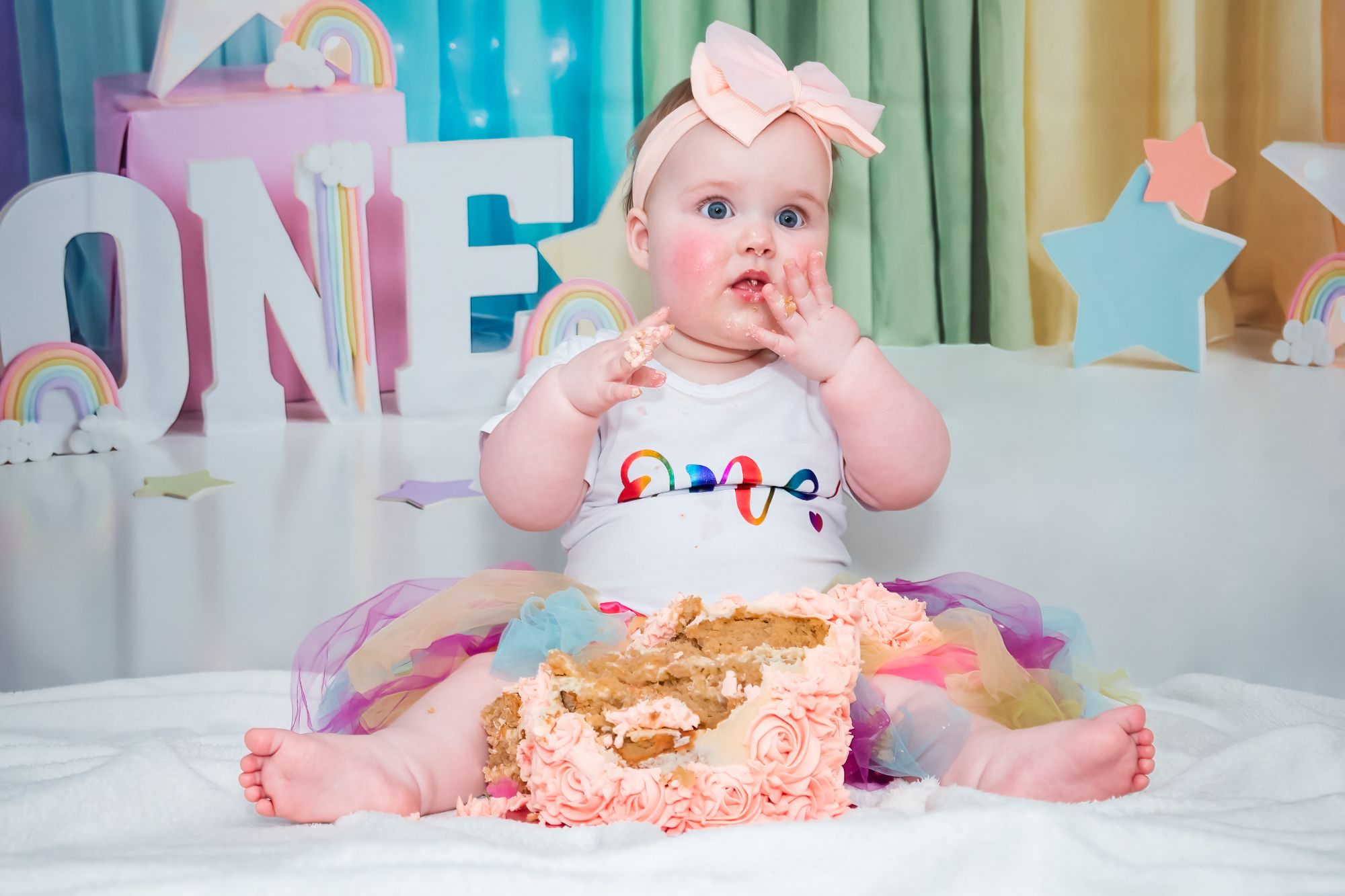 A little girl sits with one hand against her cheeck and the other hovering over her partly smashed cake. Her expression seems to be "oh my gosh is this all for me!"