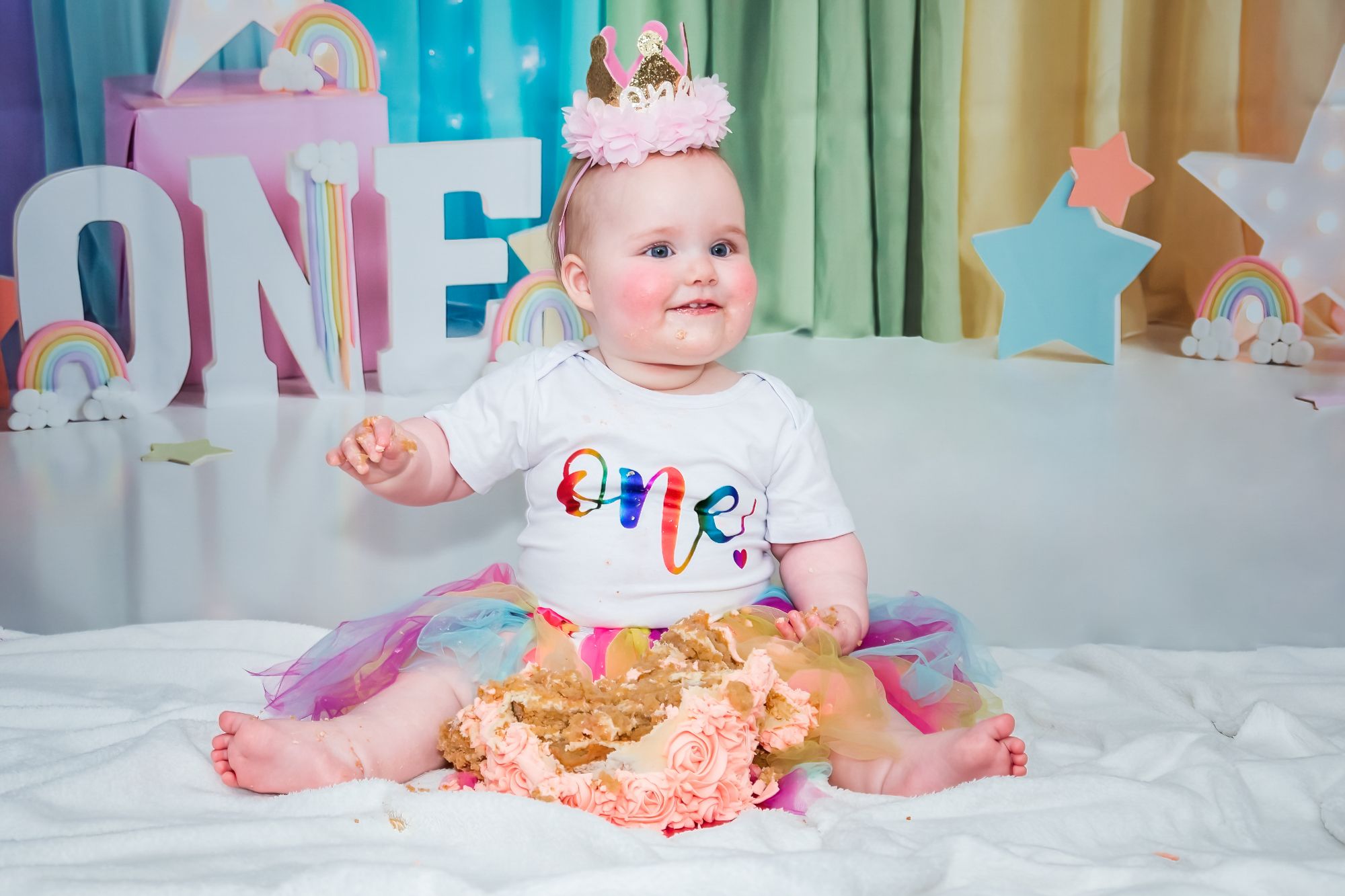 A little girl sits and smiles to her mummy who's off camera. The little girl has a mess of cake in front of her