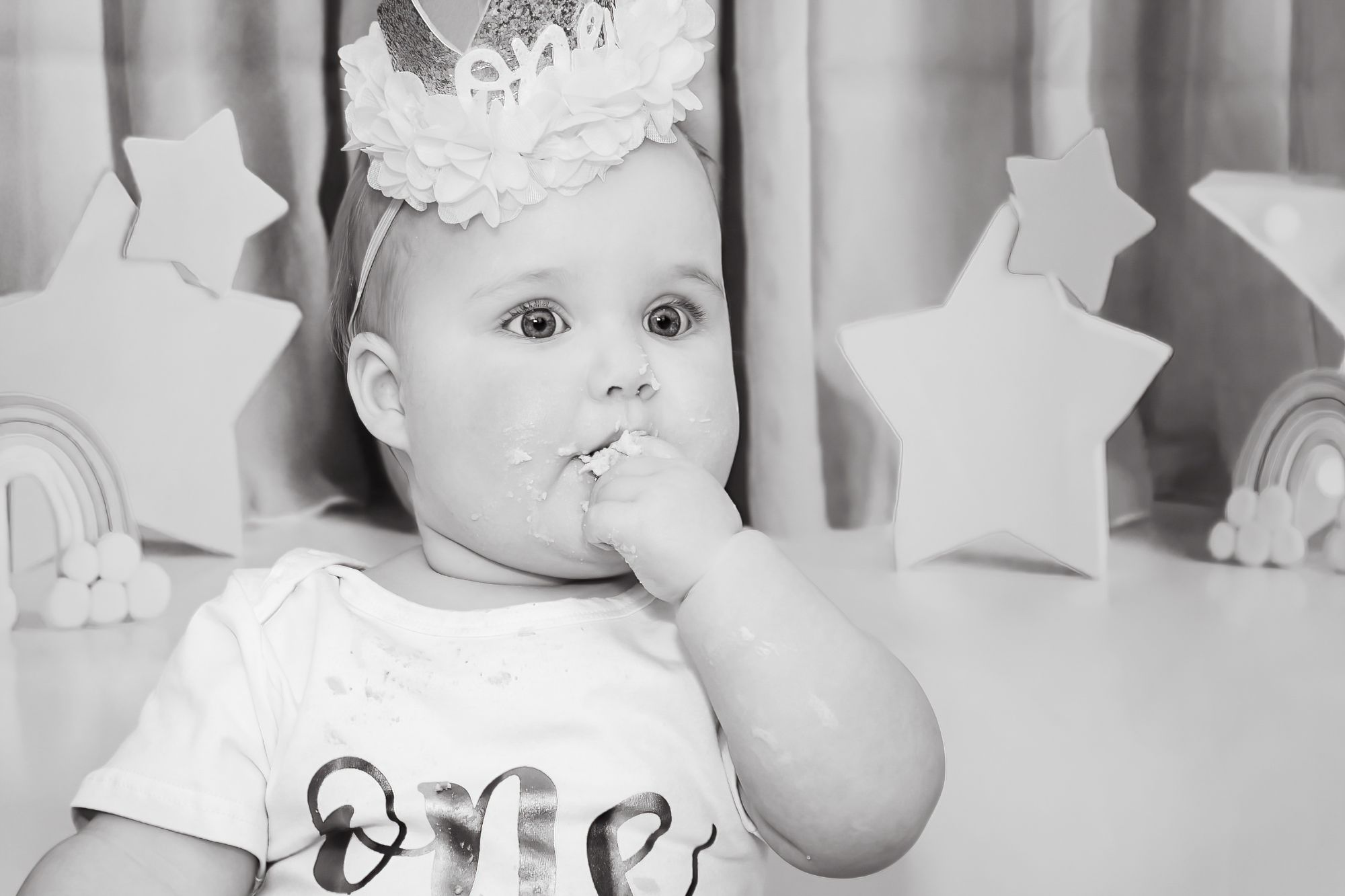A stunning black and white photograph of a little girl lost in a moment of her won during her cake smash session. She has icing on her cheek and her hand in her mouth.