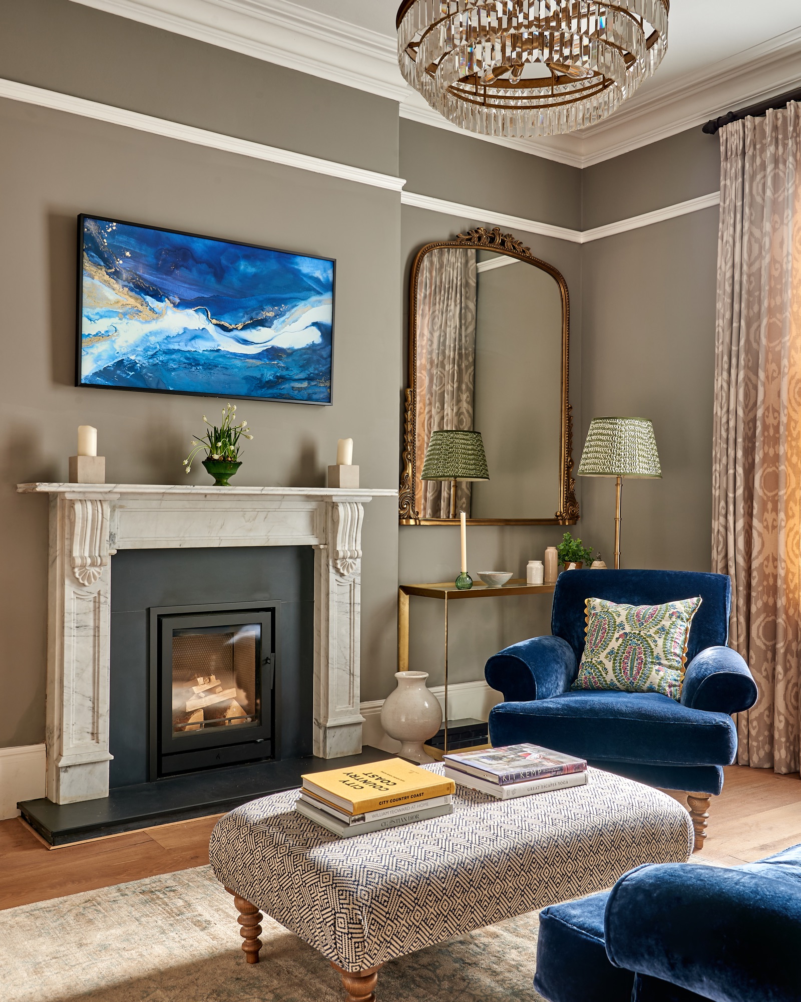 Georgian town house living area, with a stone fireplace and a Samsung frame with artwork on. Rich Blue velvet armchair from sofa.com with a wicklewood cushion, anthropologie glass consoles and large mirror,  Pooky standing lamp and a footstool  upholstered in Linwood fabric.