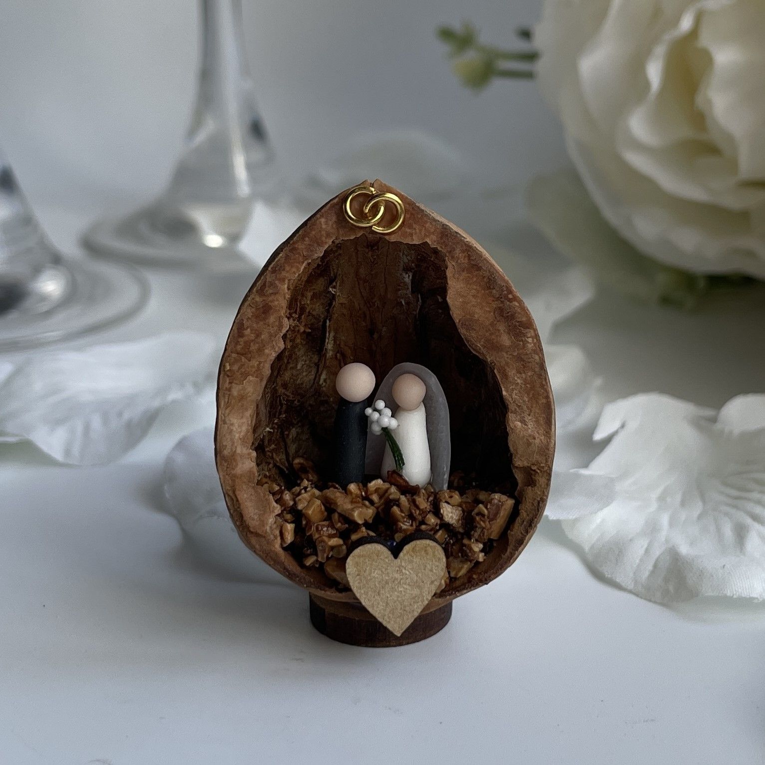 Image shows a walnut shell decoration on a table with flowers and champagne flutes. The decoration contains tiny figures of a bride and groom with two gold rings at the top and a wooden heart at the base.