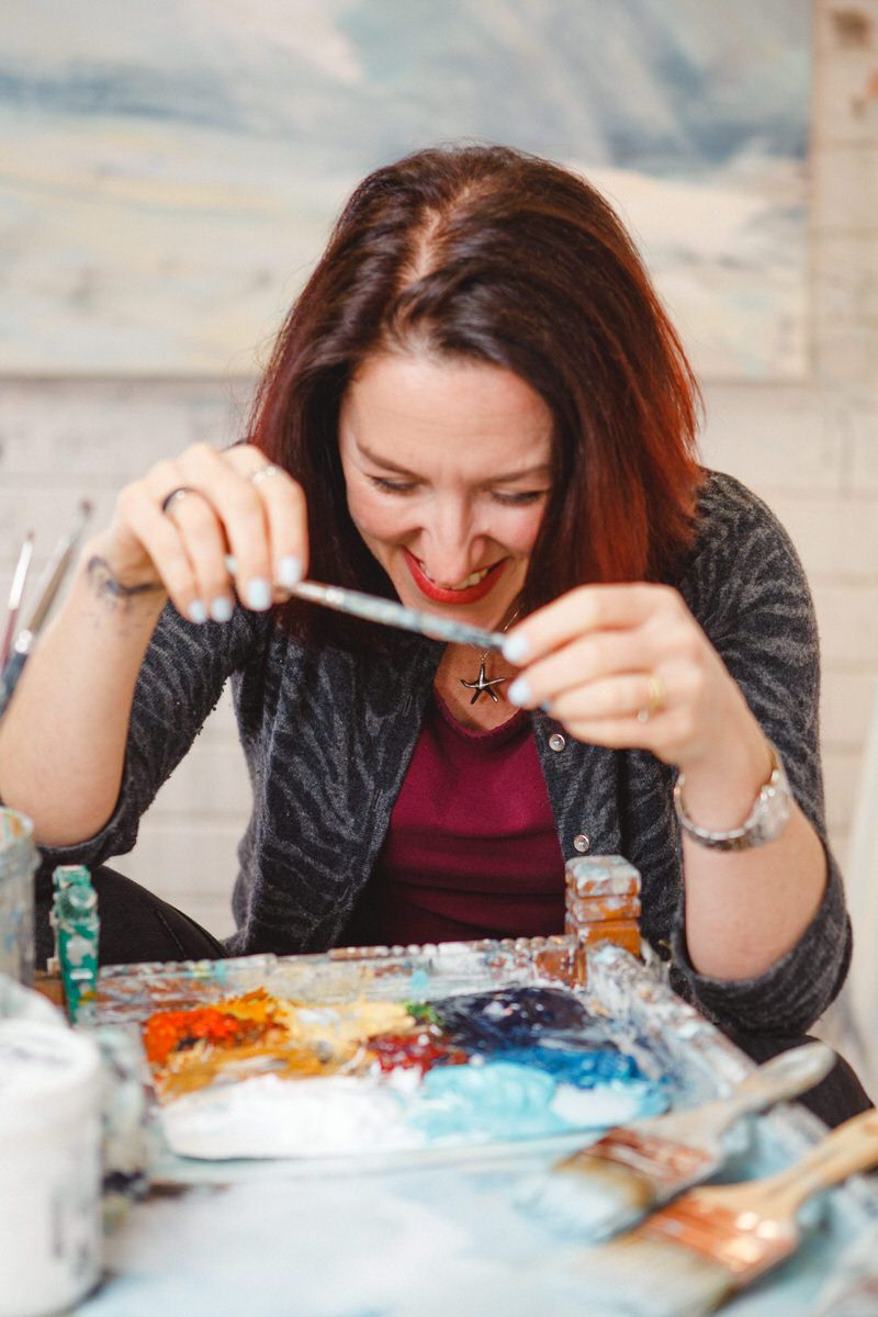 Alison laughing holding paintbrush in front of palette of colours