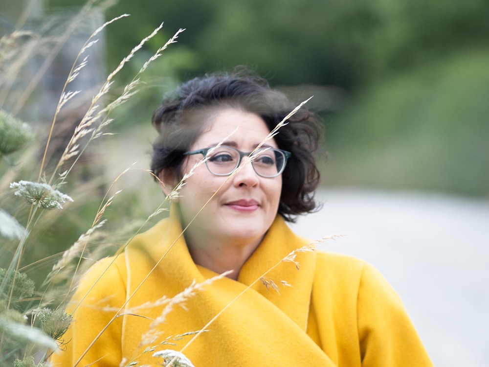 Short haired brunette woman wearing green framed glasses, mustard coloured fleece jacket and a wry smile sits outside, grasses can be seen slightly out of focus