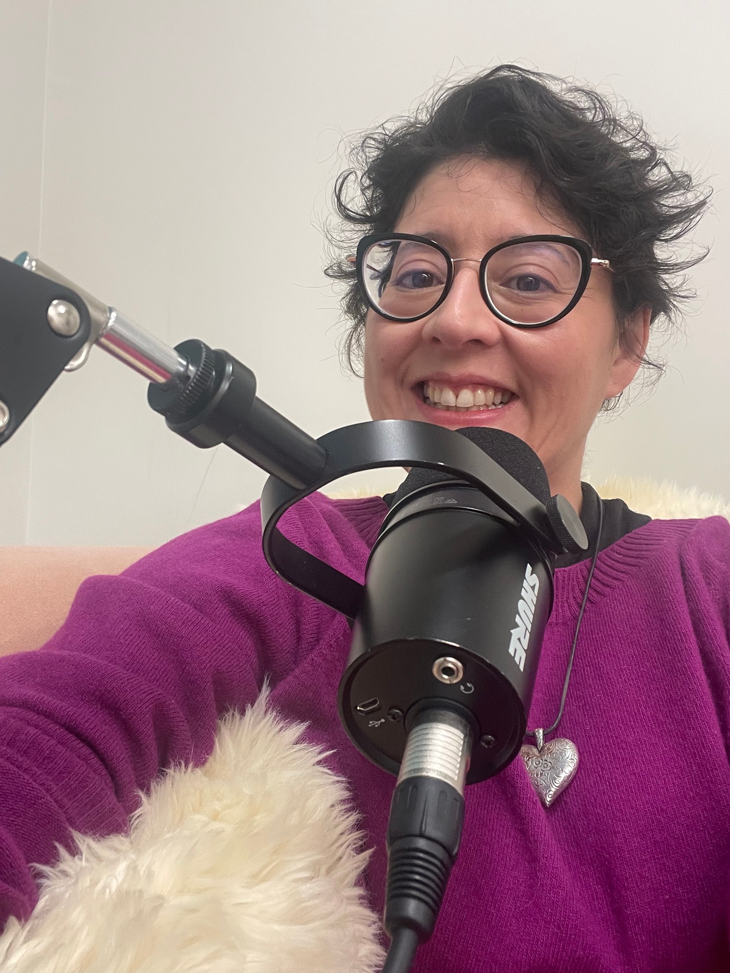 Short haired brunette woman wearing black rimmed glasses and pagenta jumper sits talking into a micriphone