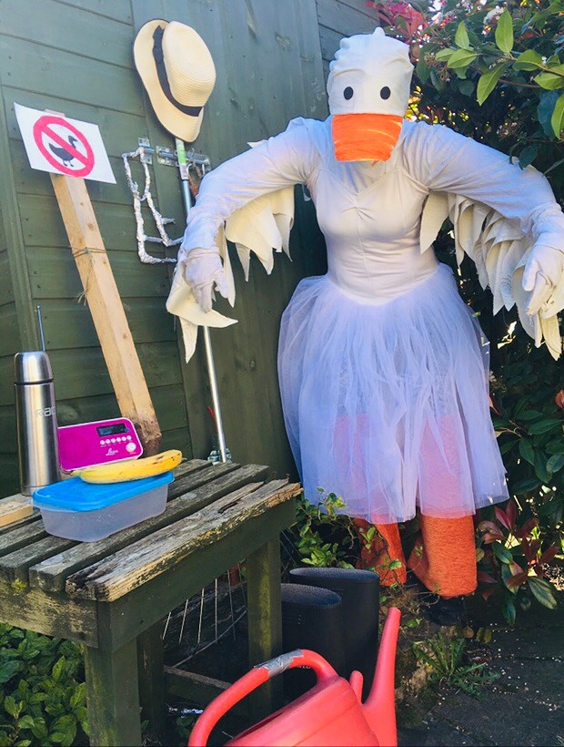 A woman is dressed up outside in the garden like a goose all in white with a fake orange beak. She stands by a dark green shed with all the elements from the game including a red watering can, radio, tea flask, lunchbox, banana, hat, rake, keys and sign post