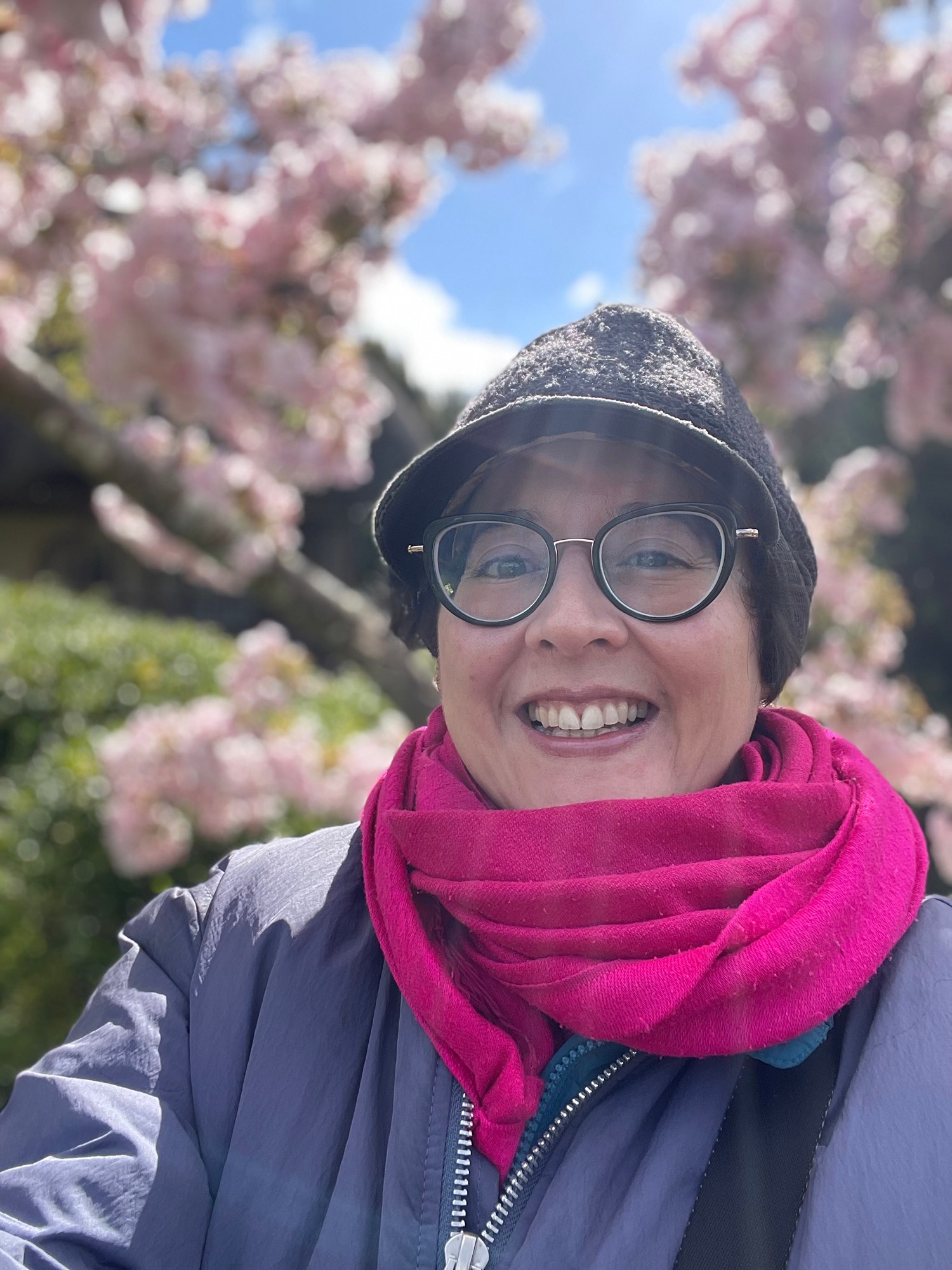 A woman smiles at the camera, she wears black rimmed glasses, a black peaked cap, magenta scarf, lilac coat and there is a magnolia in flower behind her
