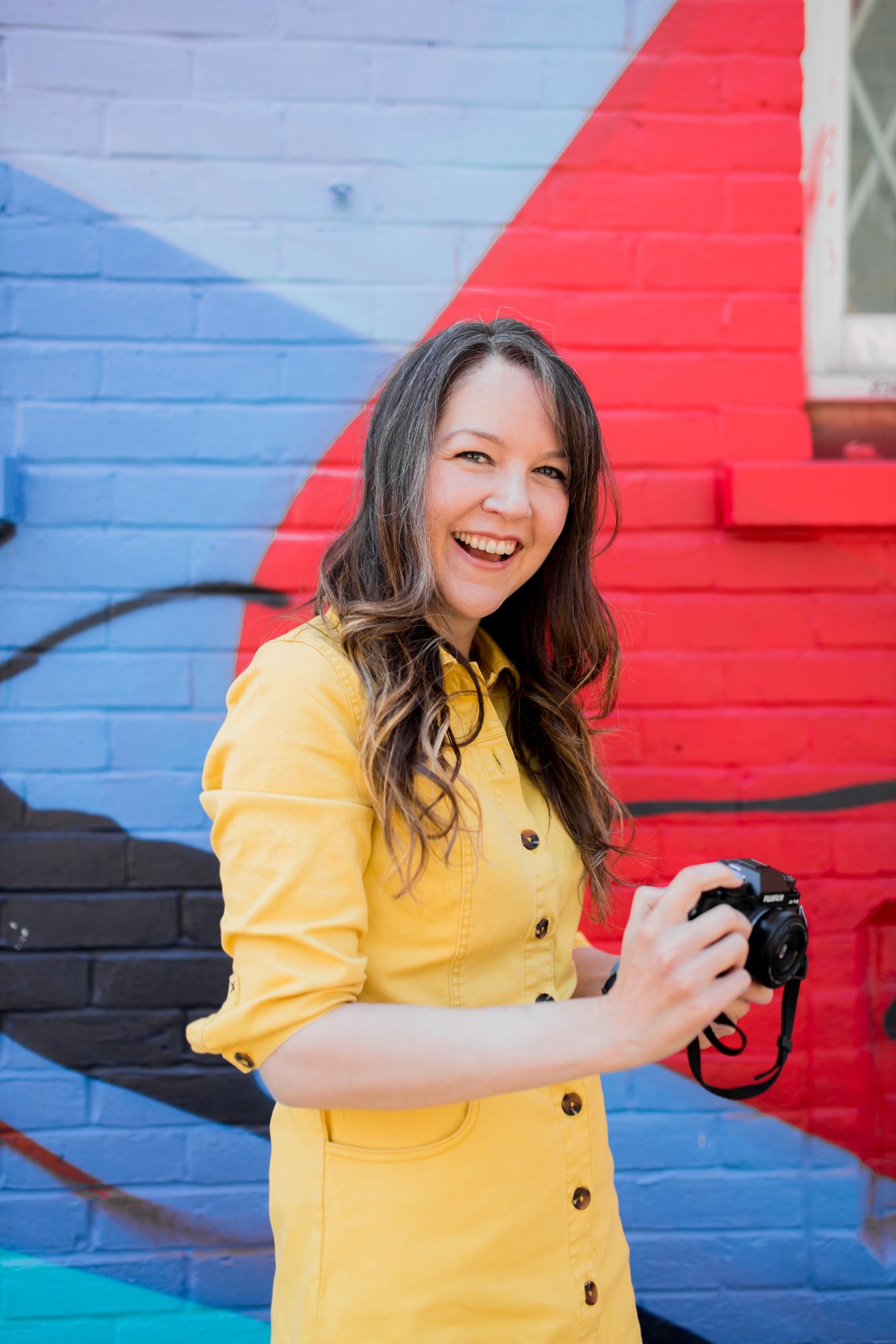 Brighton headshot photographer Lauren Psyk standing in front of a red and blue wall in a yellow dress holding her camera