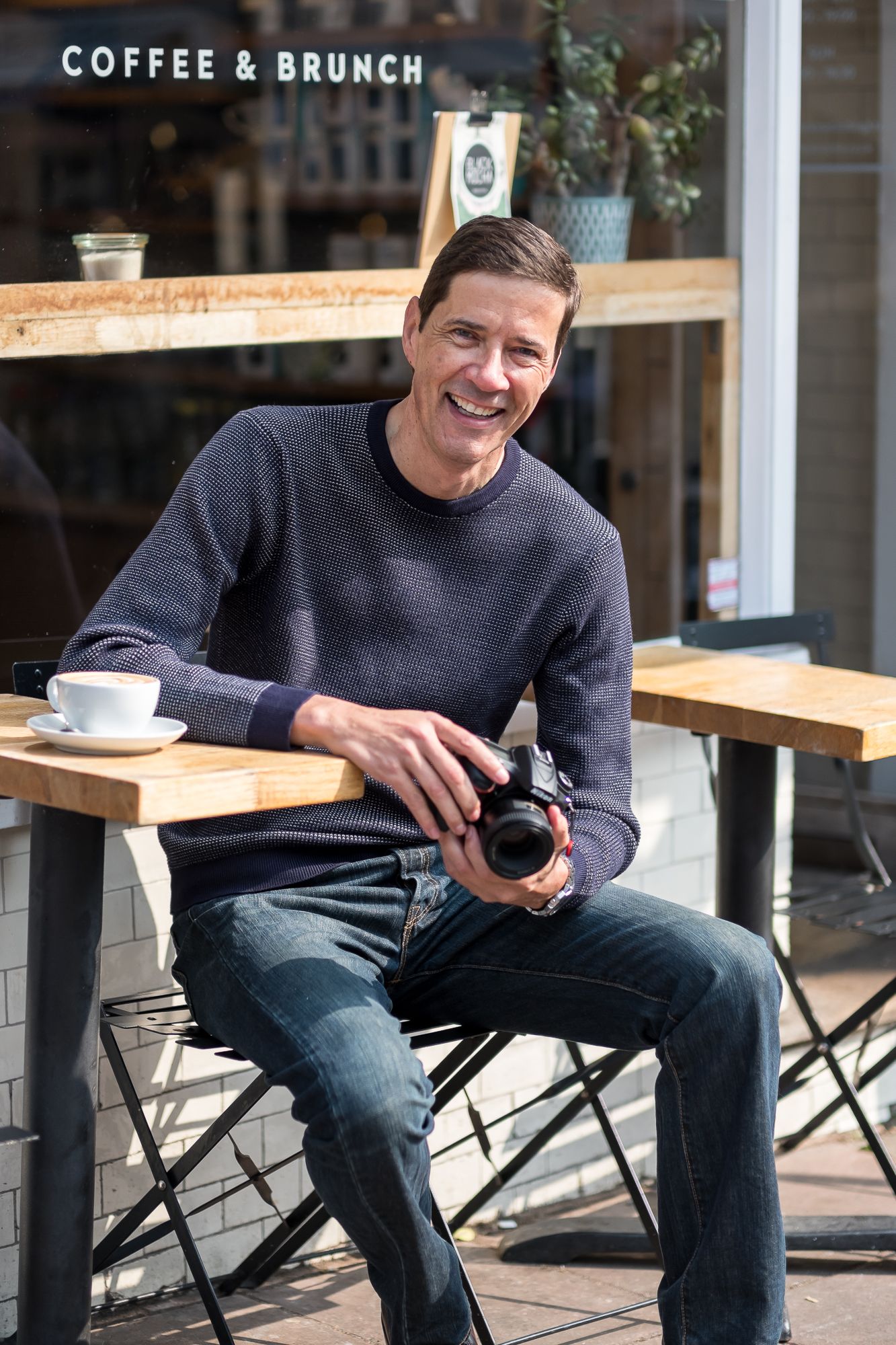 Man sitting outside Black Mocha cafe in Brighton with a cup of coffee, holding his camera and smiling