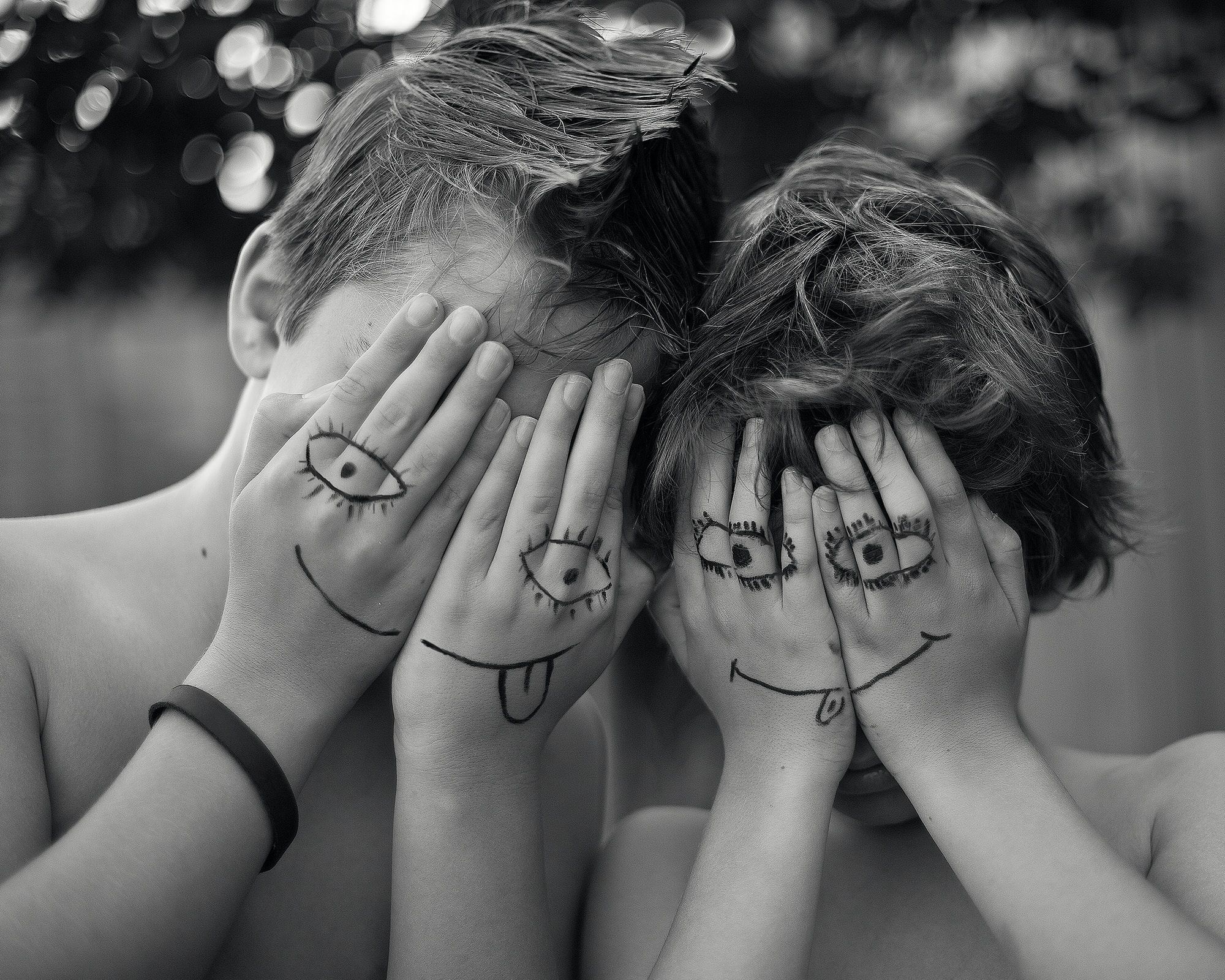two young boys are covering their faces with their hands. they have smiley faces drawn in black pen on the backs of their hands