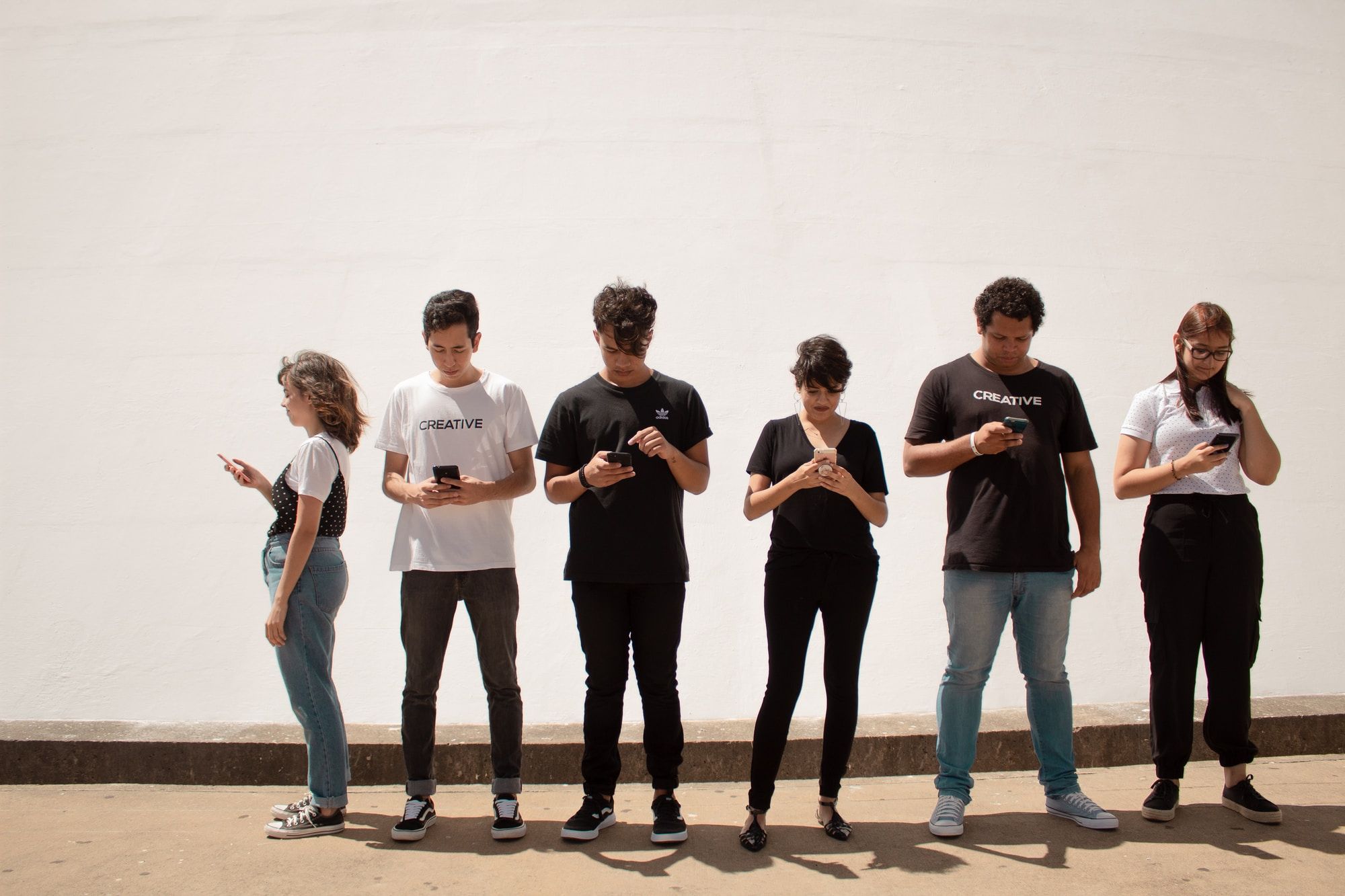 a group of teenagers stand in a line, they are all staring at their mobile phone